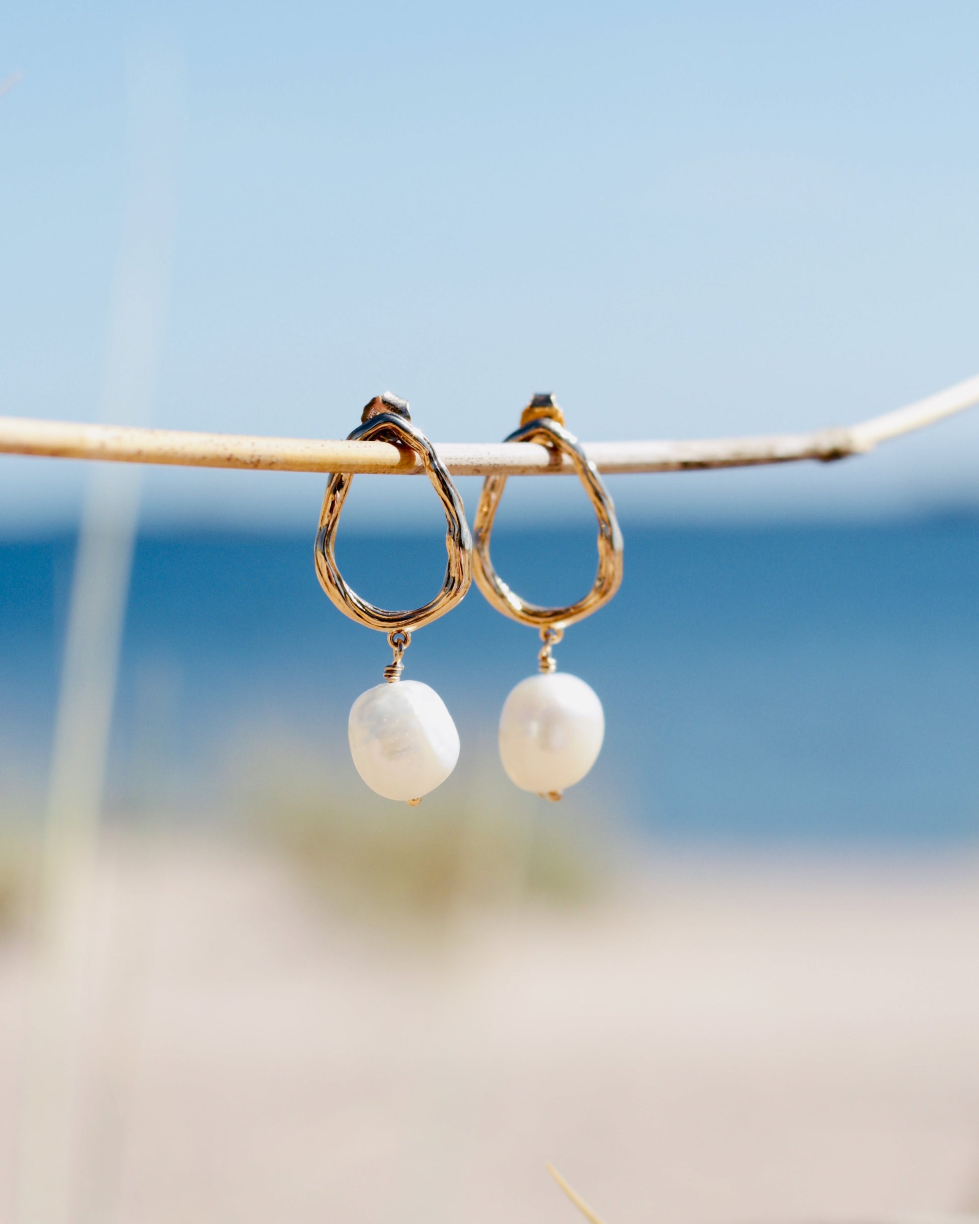 A pair of gold hoop earrings with white baroque pearl drops hanging from a thin branch outdoors with a blurred beach and blue sky background in Hanko city, Finland.