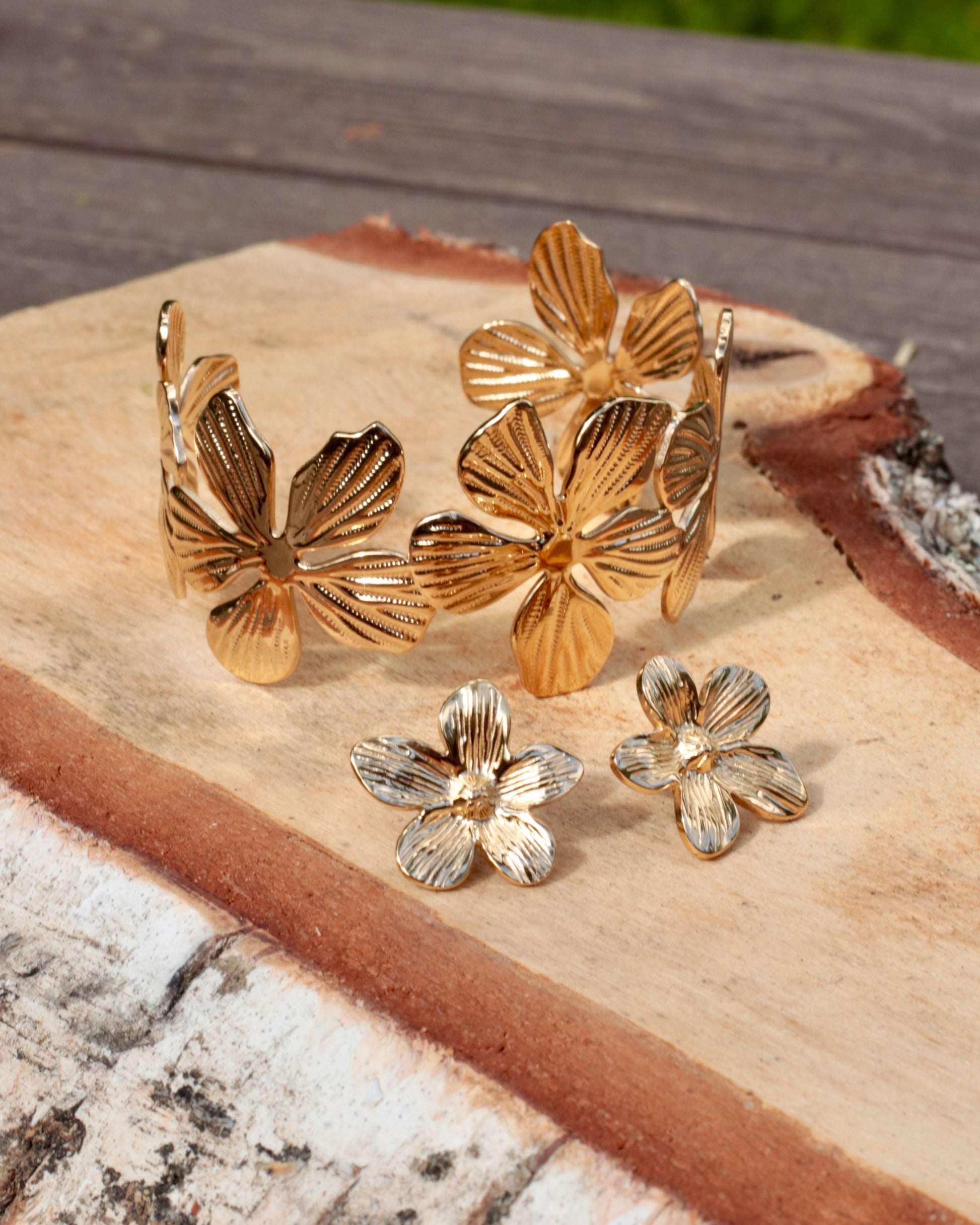 Gold-tone flower-themed jewelry set including a cuff bracelet, stud earrings, and a ring, displayed on a rustic wood slice outdoors.