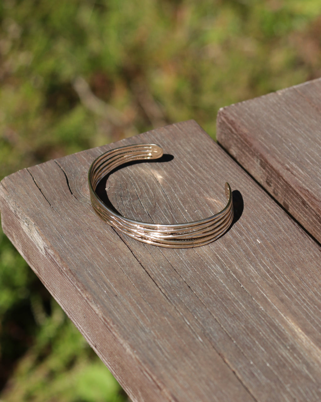 A thin, multi-strand open cuff bracelet in a warm gold tone, resting on a weathered wooden surface with a blurred green outdoor background.