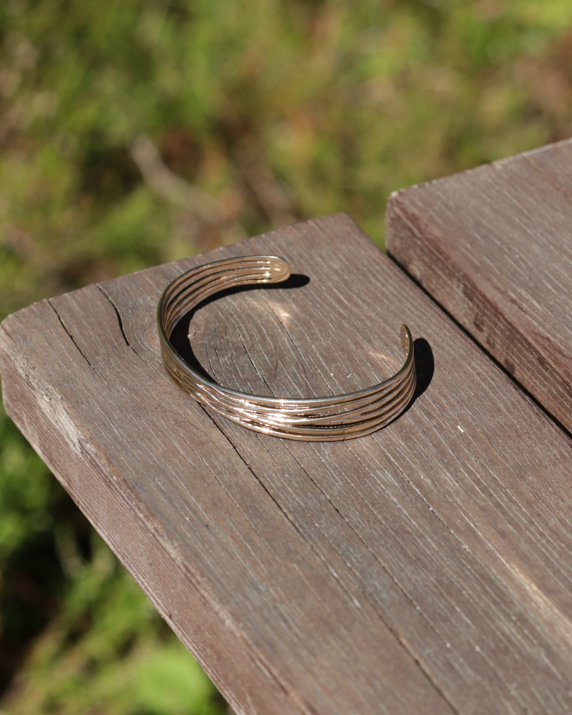 A thin, multi-strand open cuff bracelet in a warm gold tone, resting on a weathered wooden surface with a blurred green outdoor background.