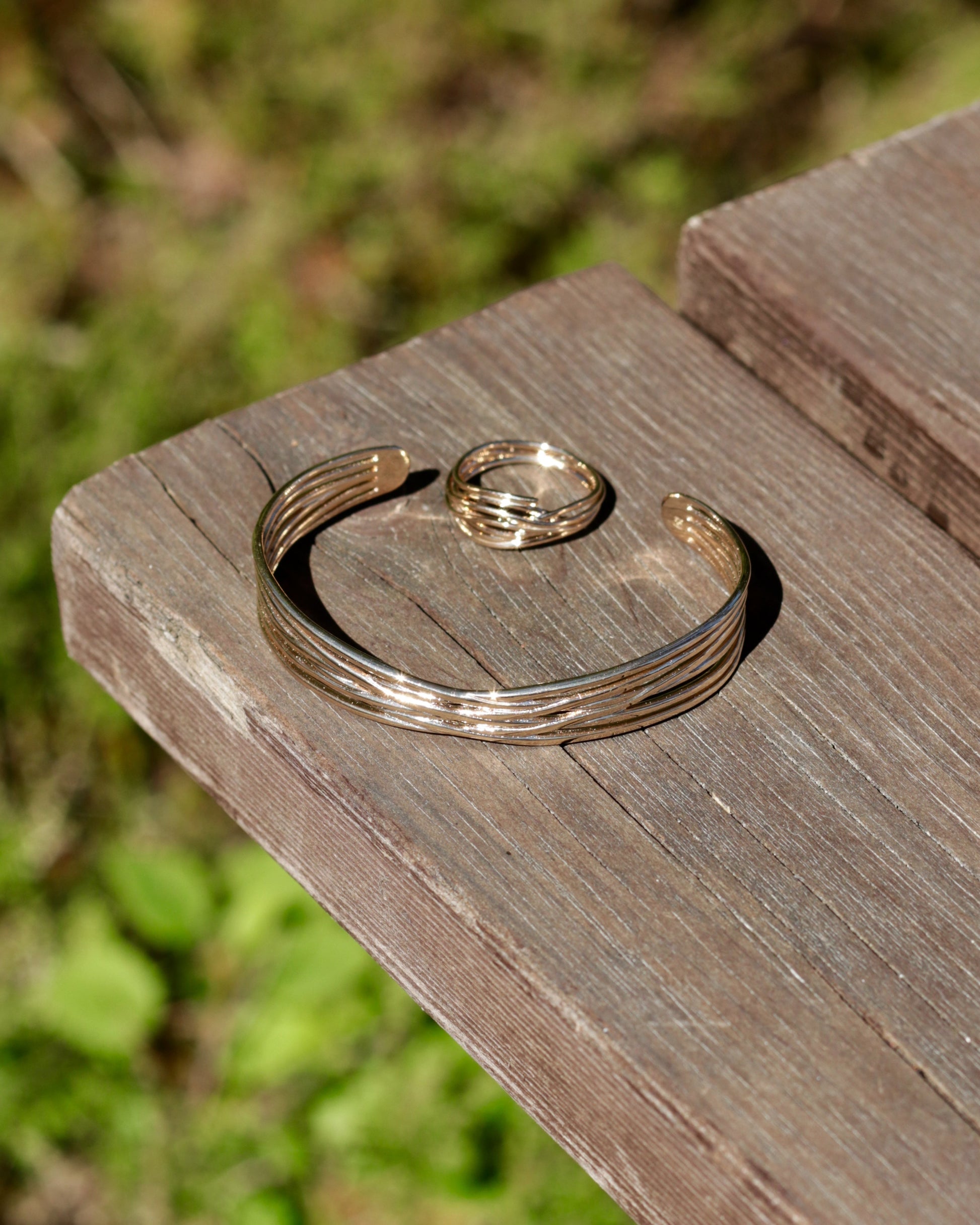 A gold-toned multi-band cuff bracelet and a matching ring displayed on a weathered wooden surface with a blurred green outdoor background.