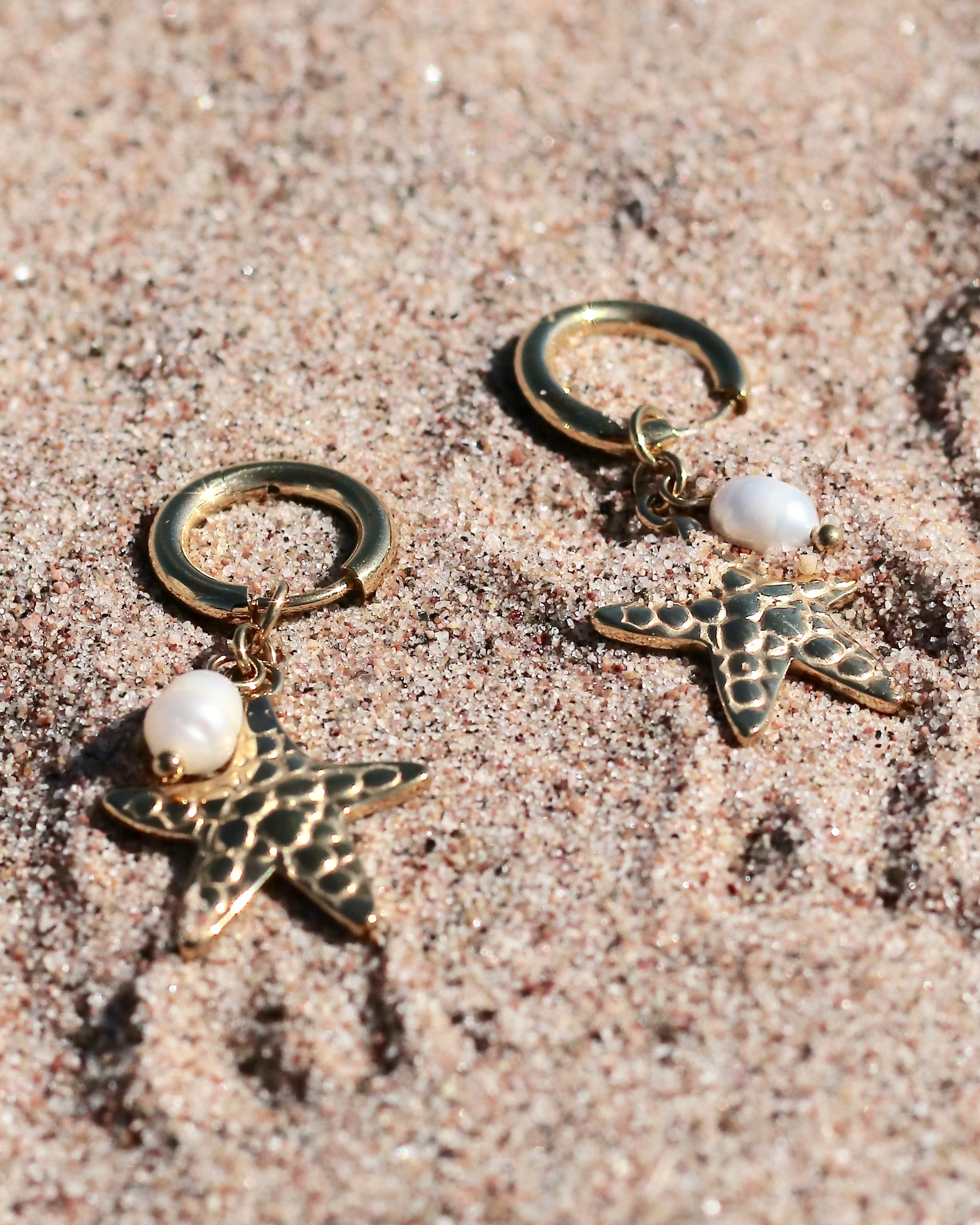 Gold-tone hoop earrings with starfish charms and freshwater pearls displayed on beach sand in Hanko, Finland