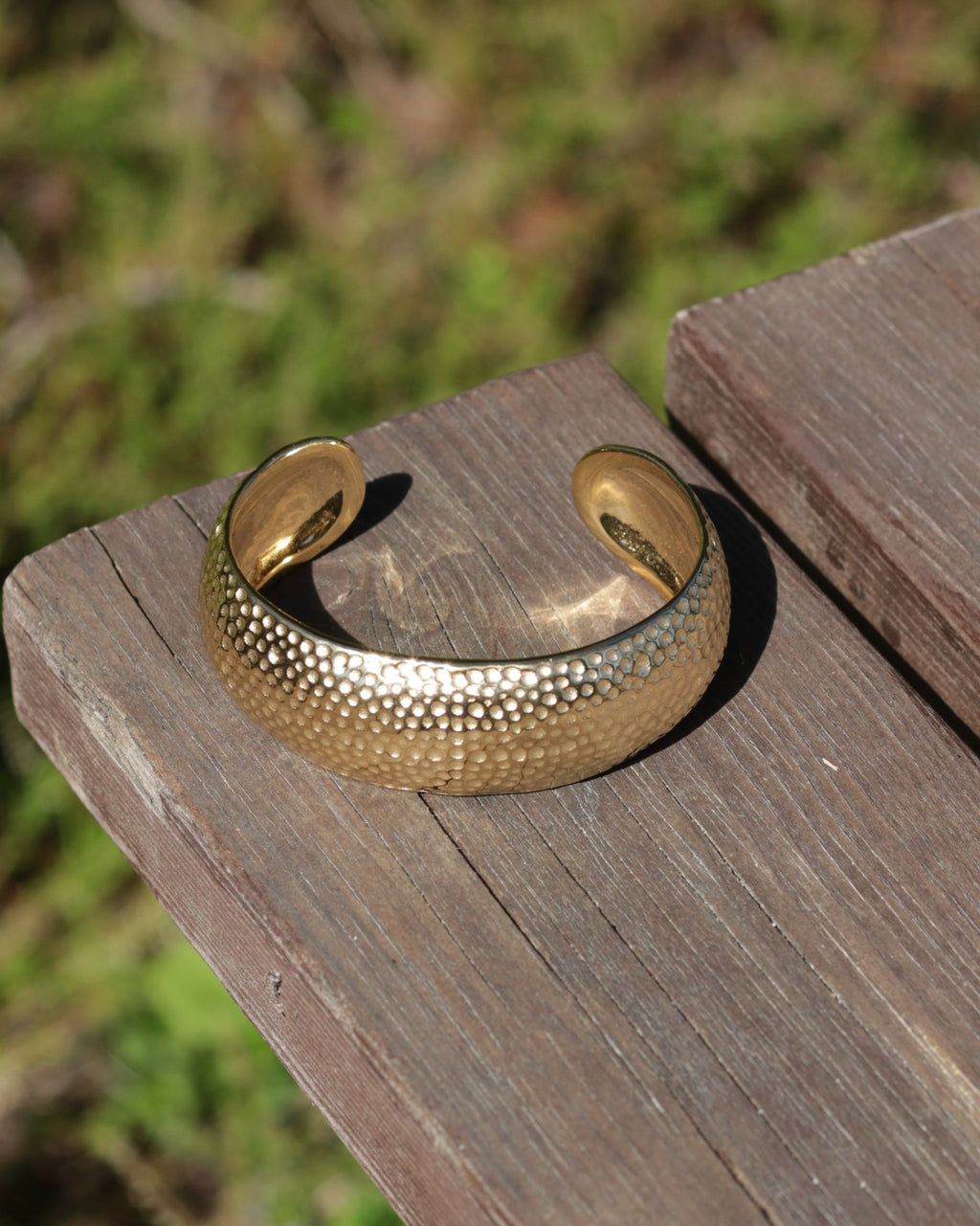 A hammered gold cuff bracelet resting on a weathered wooden board with a blurred green outdoor background.