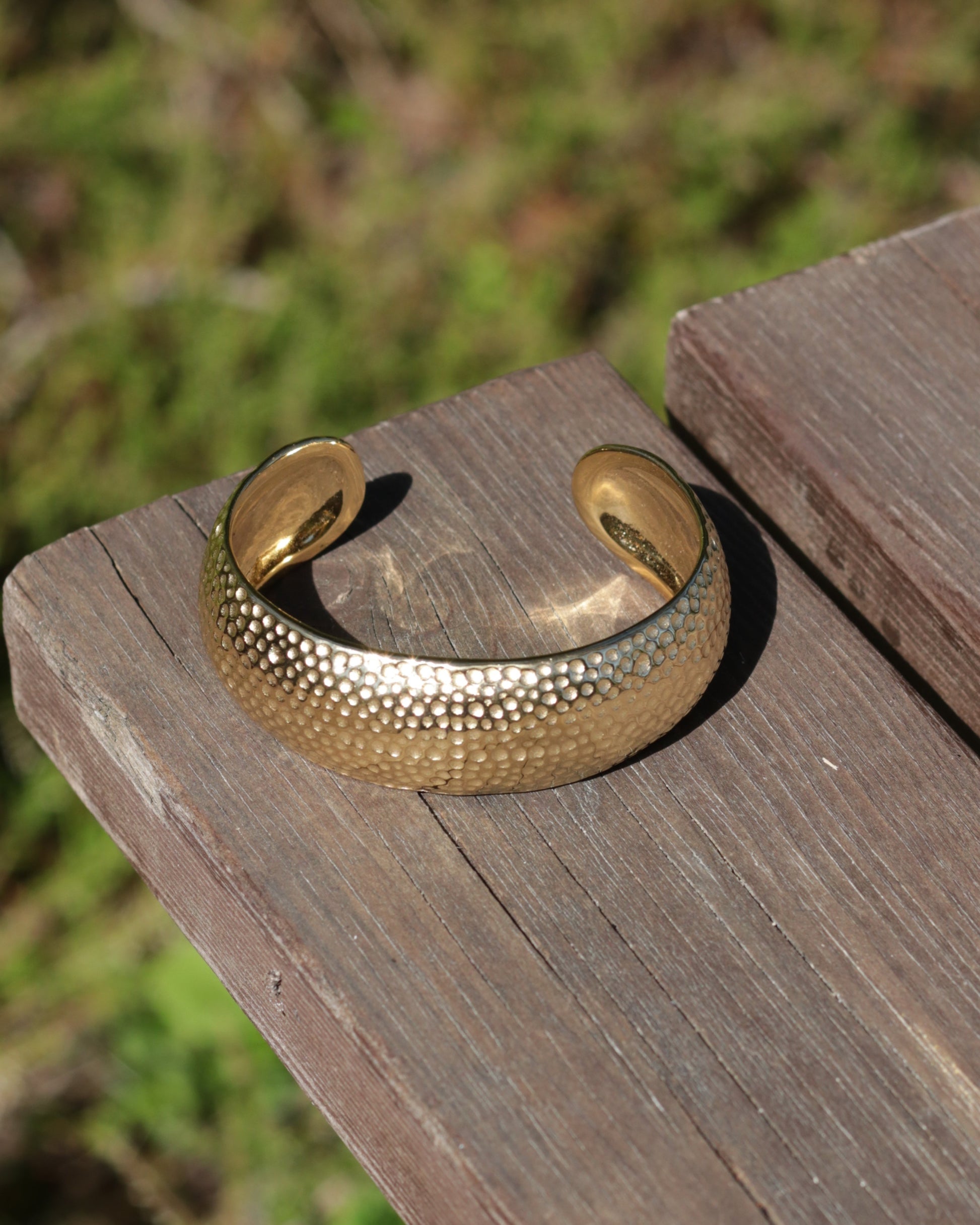 A hammered gold cuff bracelet resting on a weathered wooden board with a blurred green outdoor background.