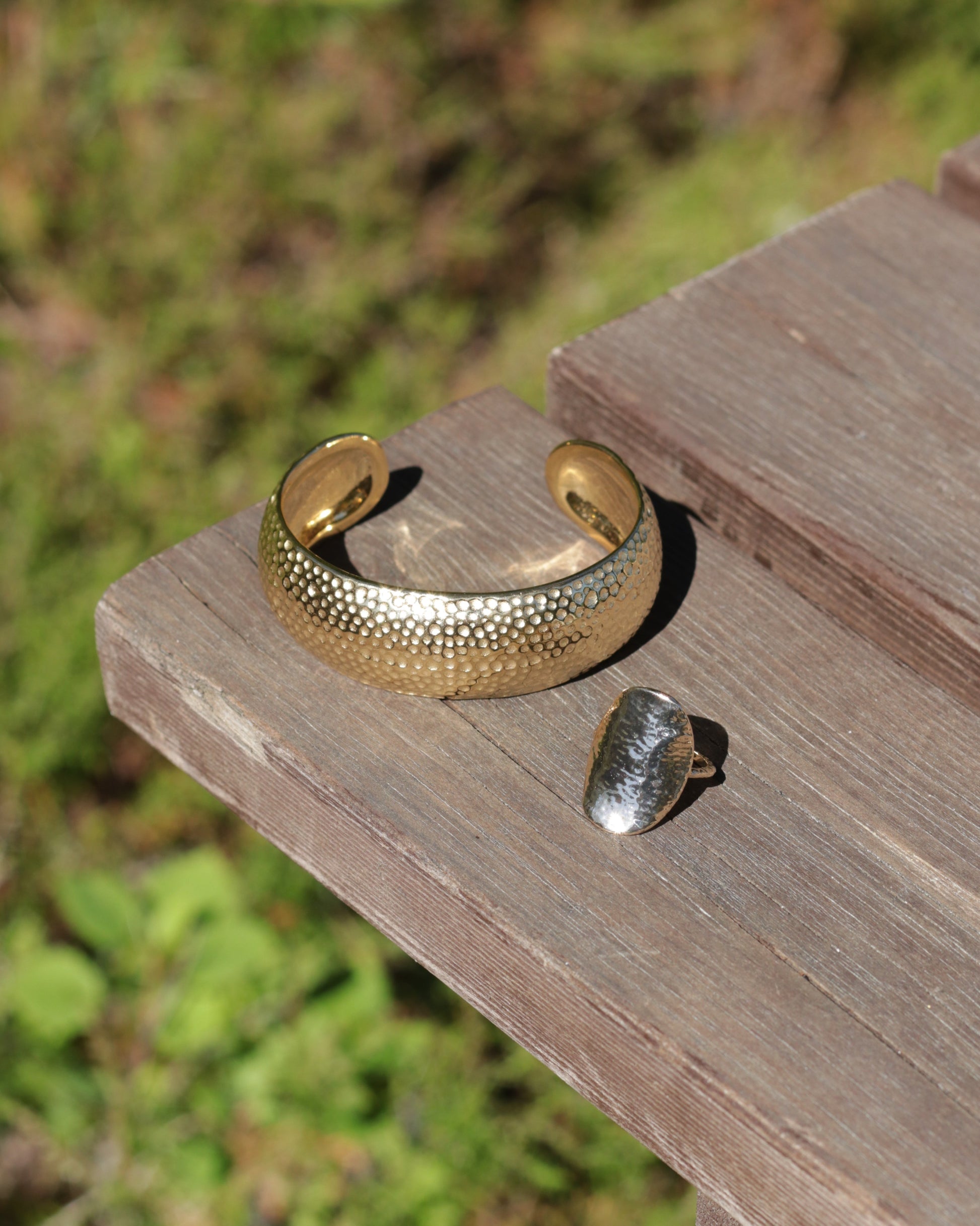 A textured gold cuff bracelet and a silver ring displayed on a weathered wooden bench with a blurred green outdoor background.