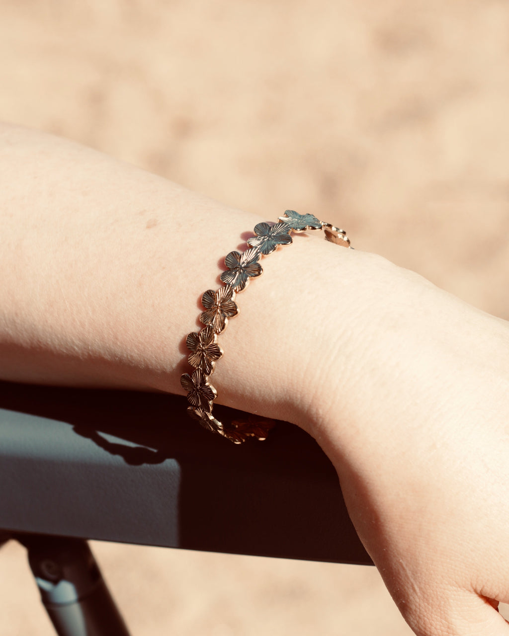 A close-up of a person's wrist wearing a delicate gold-tone bracelet with small flower or leaf motifs, photographed outdoors against a warm sandy background.