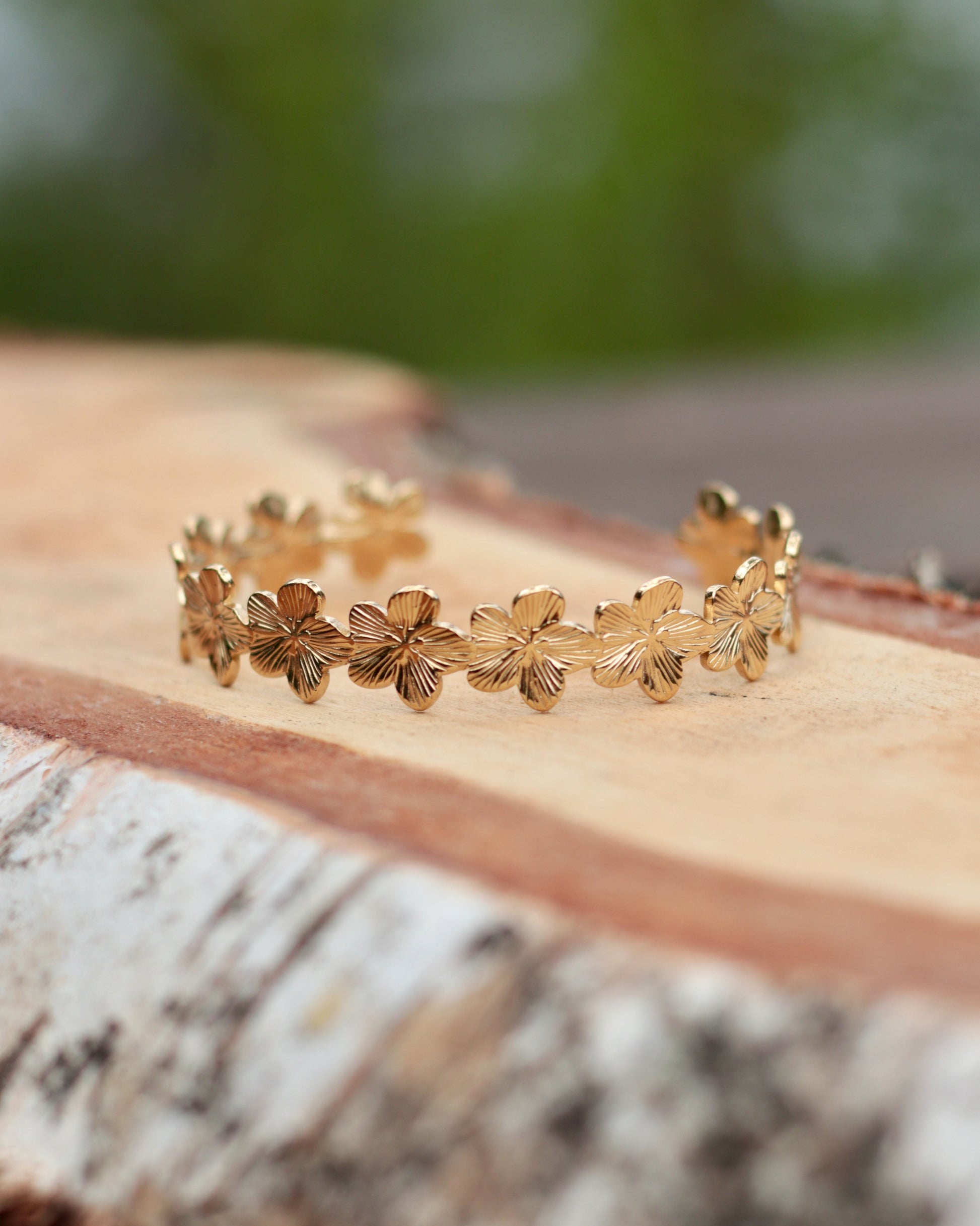 A delicate gold bangle bracelet featuring a continuous row of small textured flower motifs, resting on a rustic piece of birch wood with a blurred green outdoor background.