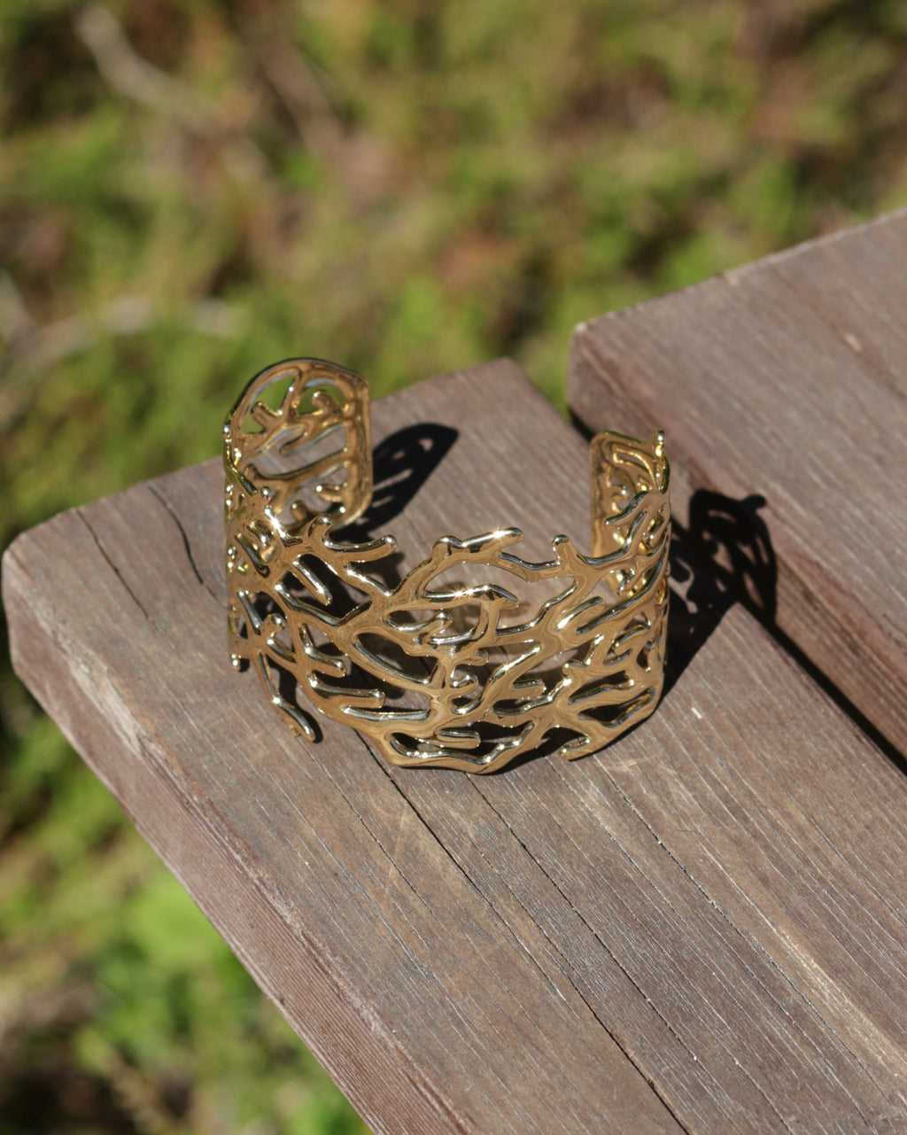 A gold-toned open cuff bracelet with an intricate, branch-like organic pattern, resting on a weathered wooden surface with a blurred green outdoor background.