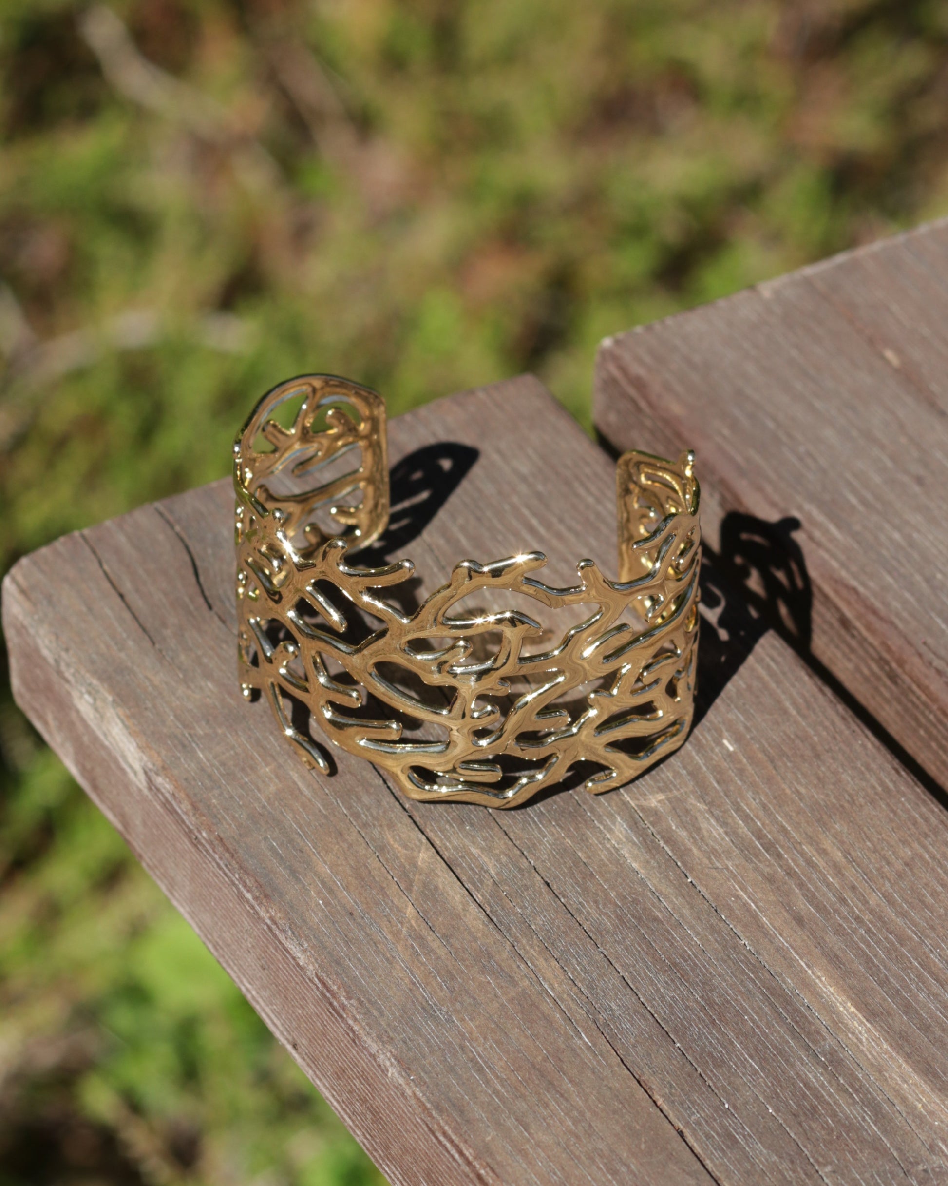 A gold-toned open cuff bracelet with an intricate, branch-like organic pattern, resting on a weathered wooden surface with a blurred green outdoor background.
