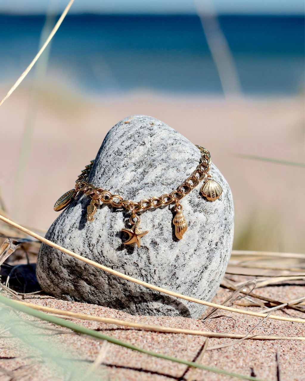 Gold charm bracelet with seashell and starfish pendants displayed on a beach stone by the sea by HNKO Finland