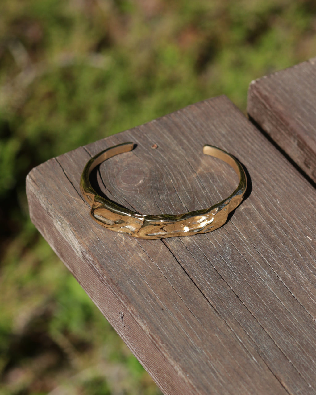 A hammered gold-tone metal cuff bracelet resting on a weathered wooden plank with a blurred green outdoor background.