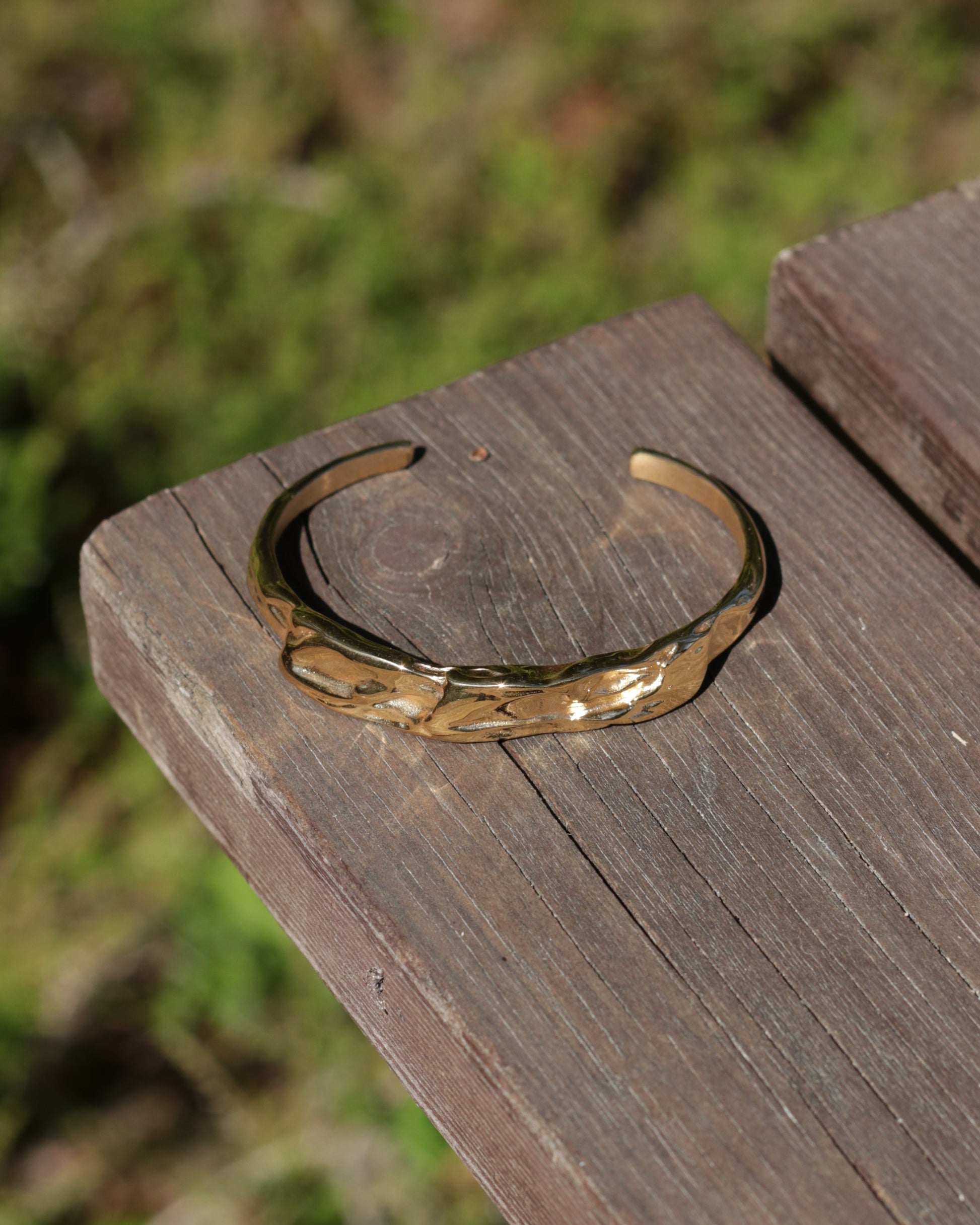 A hammered gold-tone metal cuff bracelet resting on a weathered wooden plank with a blurred green outdoor background.