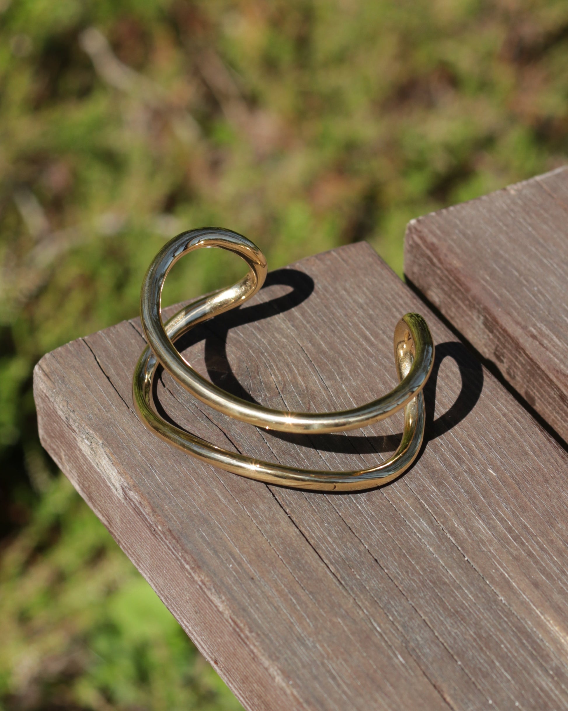 A polished gold-tone metal cuff bracelet with a twisted, open design, resting on a weathered wooden surface with a blurred green outdoor background.