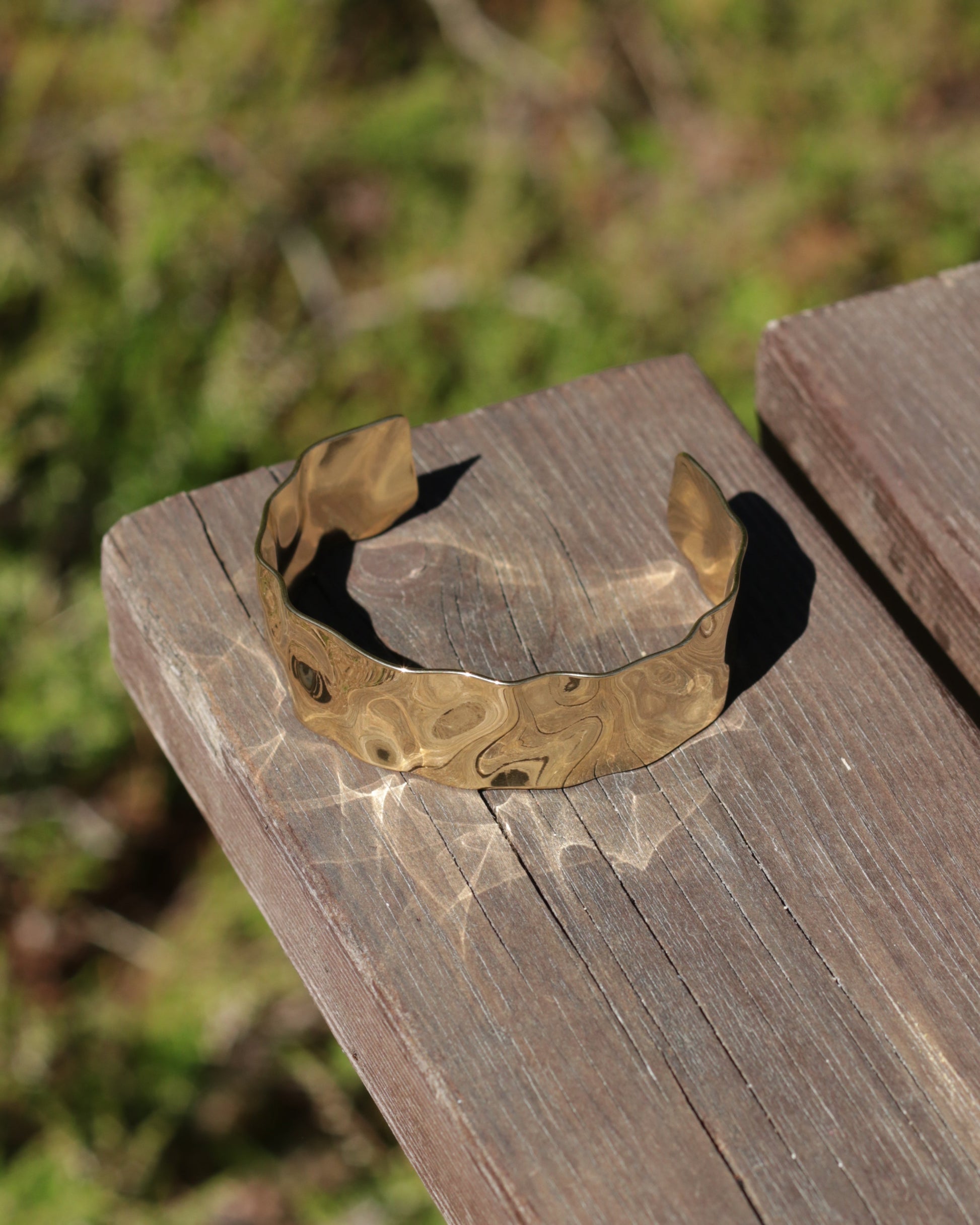 A hammered metallic cuff bracelet with a wavy, organic pattern in gold and bronze tones, resting on a weathered wooden plank with a blurred outdoor background.