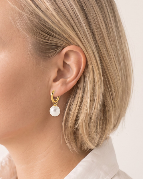 A close-up side profile of a woman with blonde hair wearing a gold drop earring with a white teardrop pearl pendant.