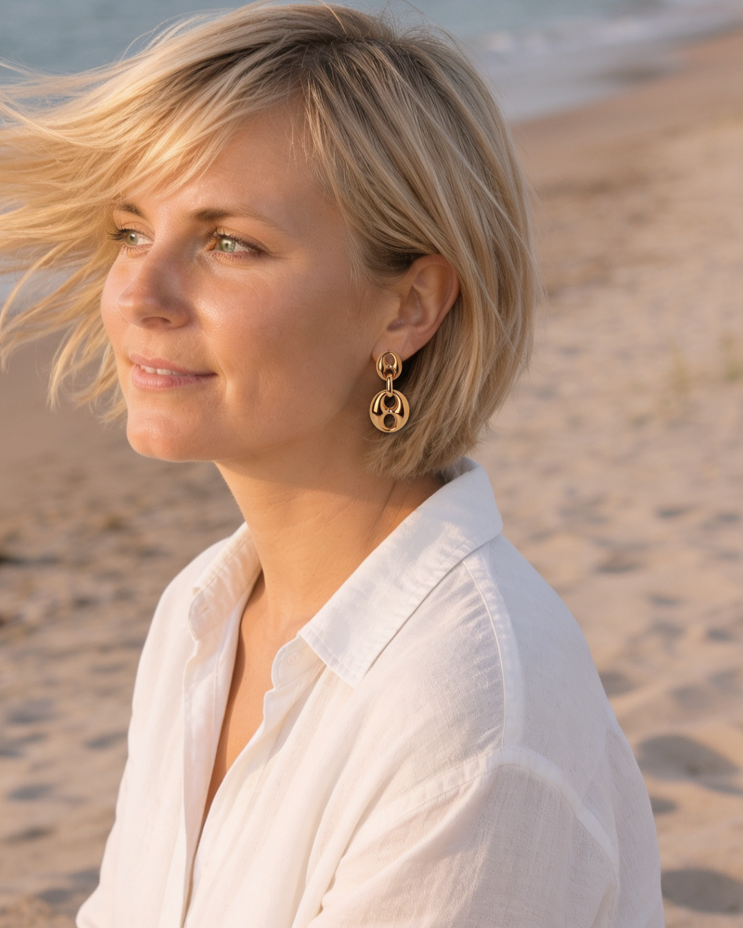 A blonde woman in a white collared shirt wearing a gold hoop earring with a circular cutout design, photographed outdoors on a sandy beach with the ocean in the background.
