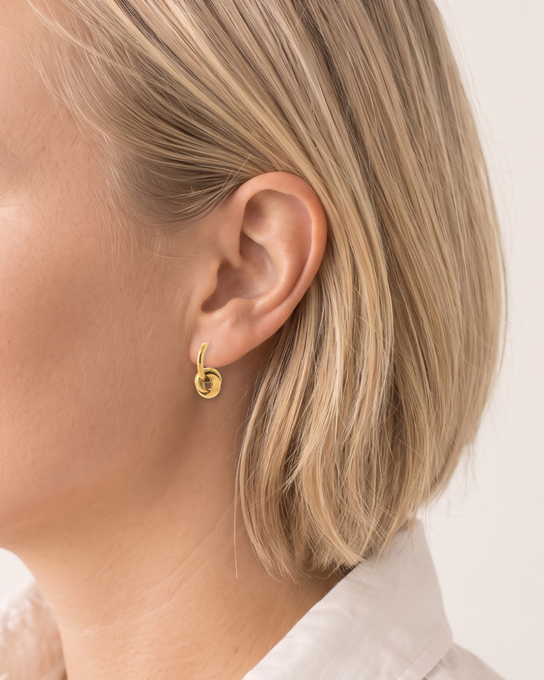 A close-up side profile of a model with blonde hair wearing a small gold hoop earring with a circular textured detail.
