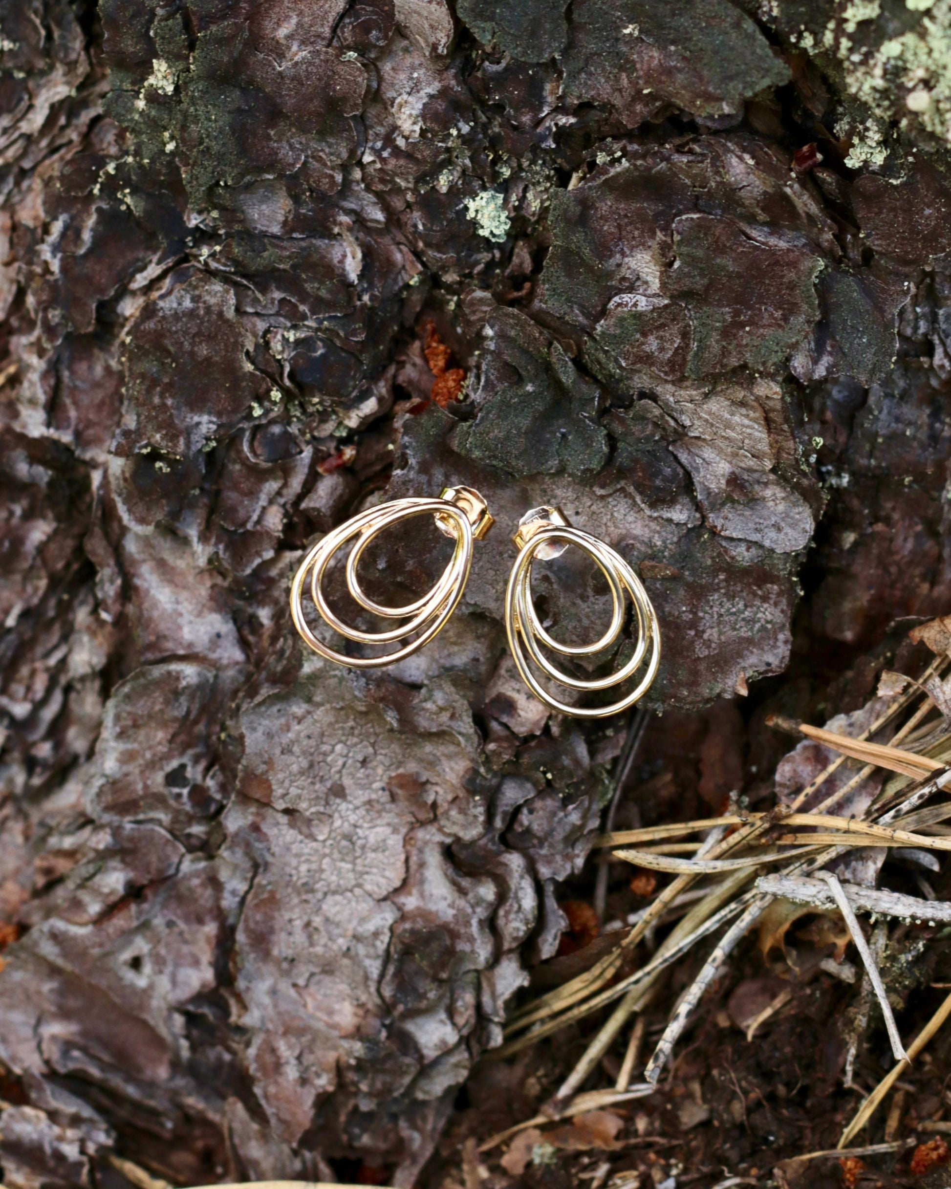 A pair of gold hoop earrings with three stacked, wavy concentric hoops, resting on textured tree bark outdoors.