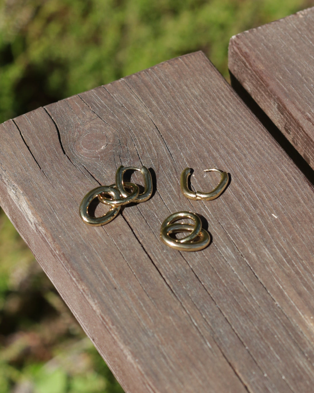 Four small gold-toned metal rings or chain-link style jewelry pieces arranged on a weathered wooden surface with blurred greenery in the background.