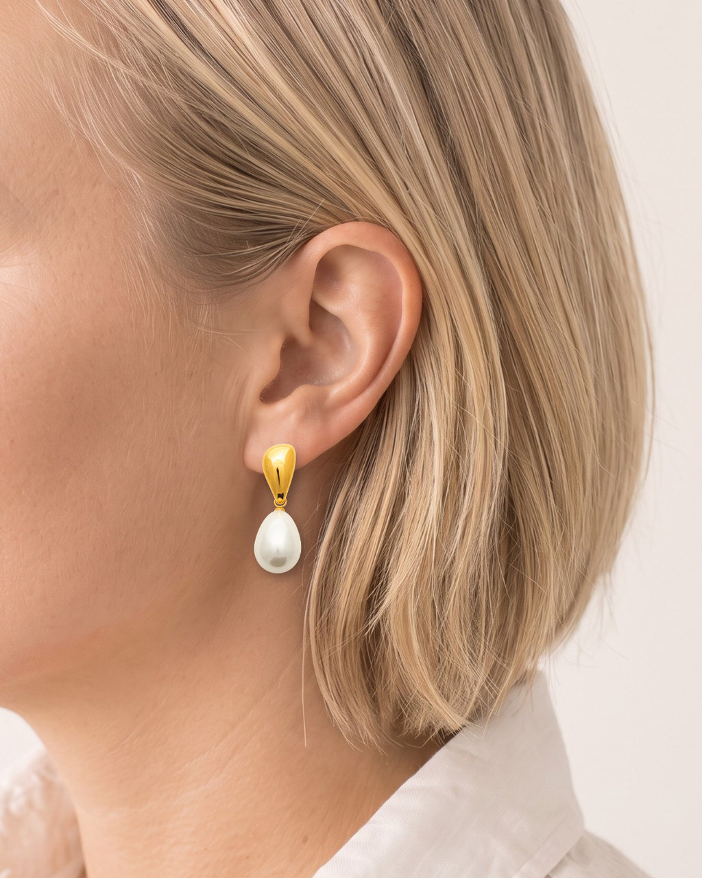 A close-up side profile of a woman with blonde hair wearing a gold drop earring with a white teardrop pearl pendant.