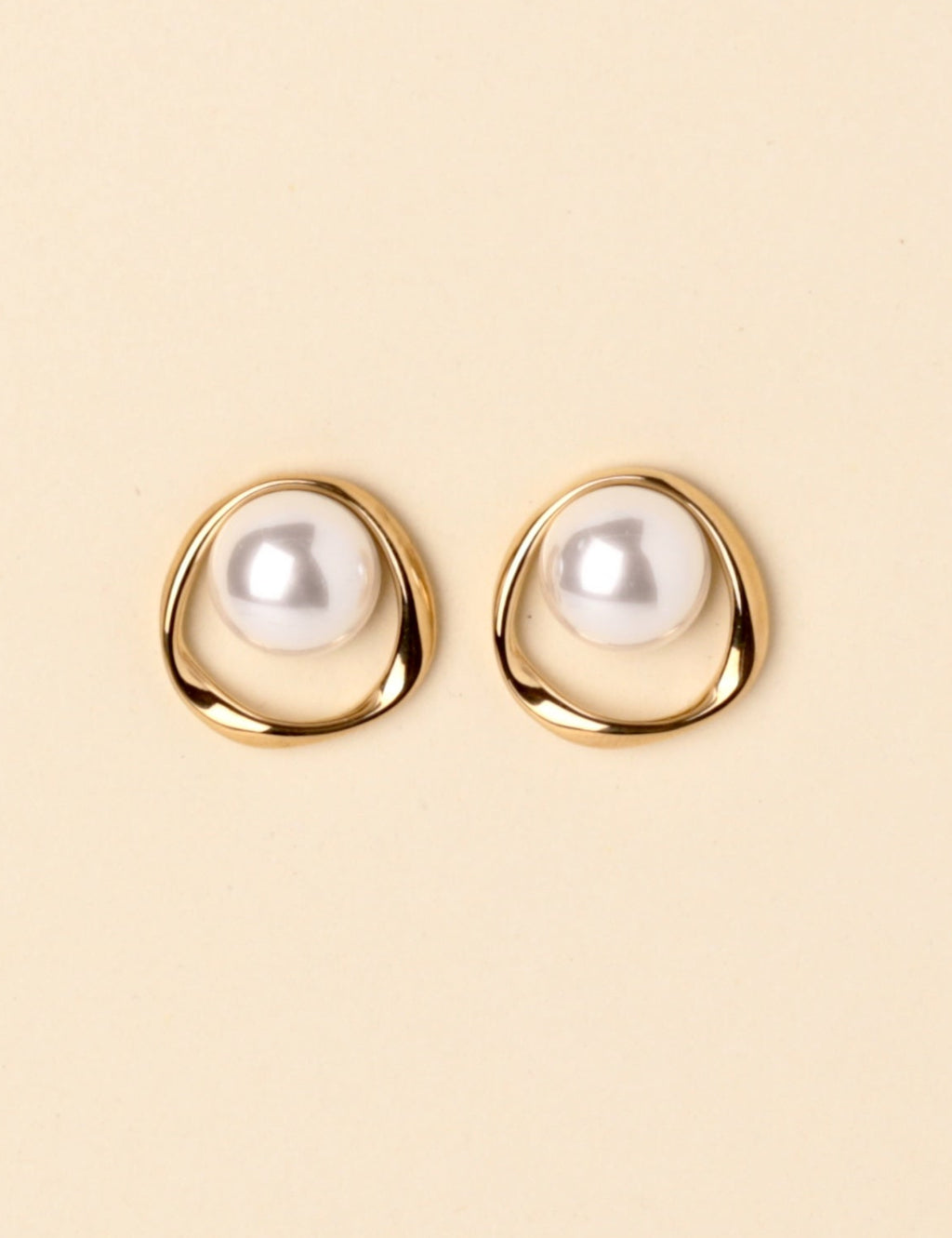 A pair of gold-toned stud earrings with a white pearl set in an organic, open circular shape, displayed on a solid light beige background.