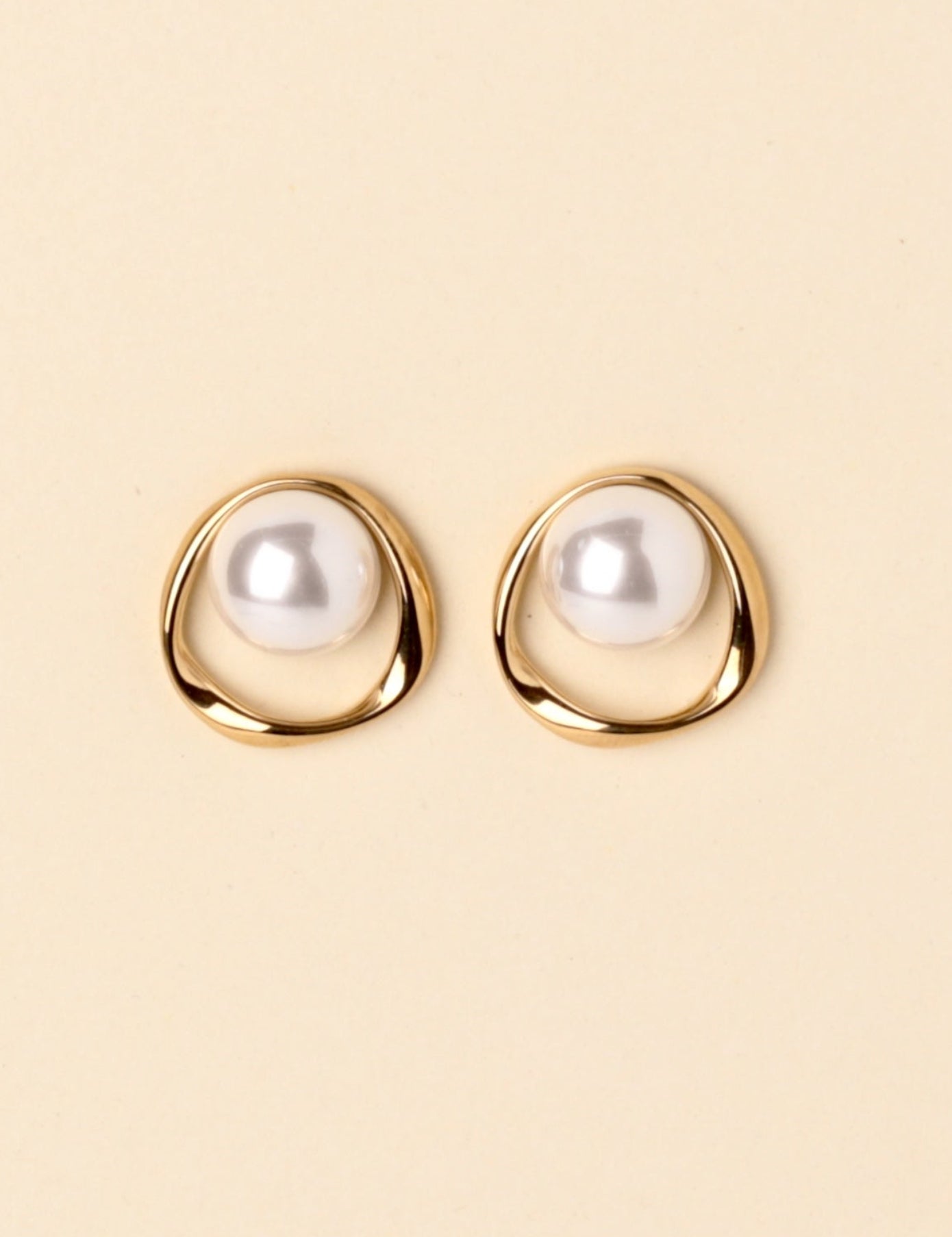 A pair of gold-toned stud earrings with a white pearl set in an organic, open circular shape, displayed on a solid light beige background.