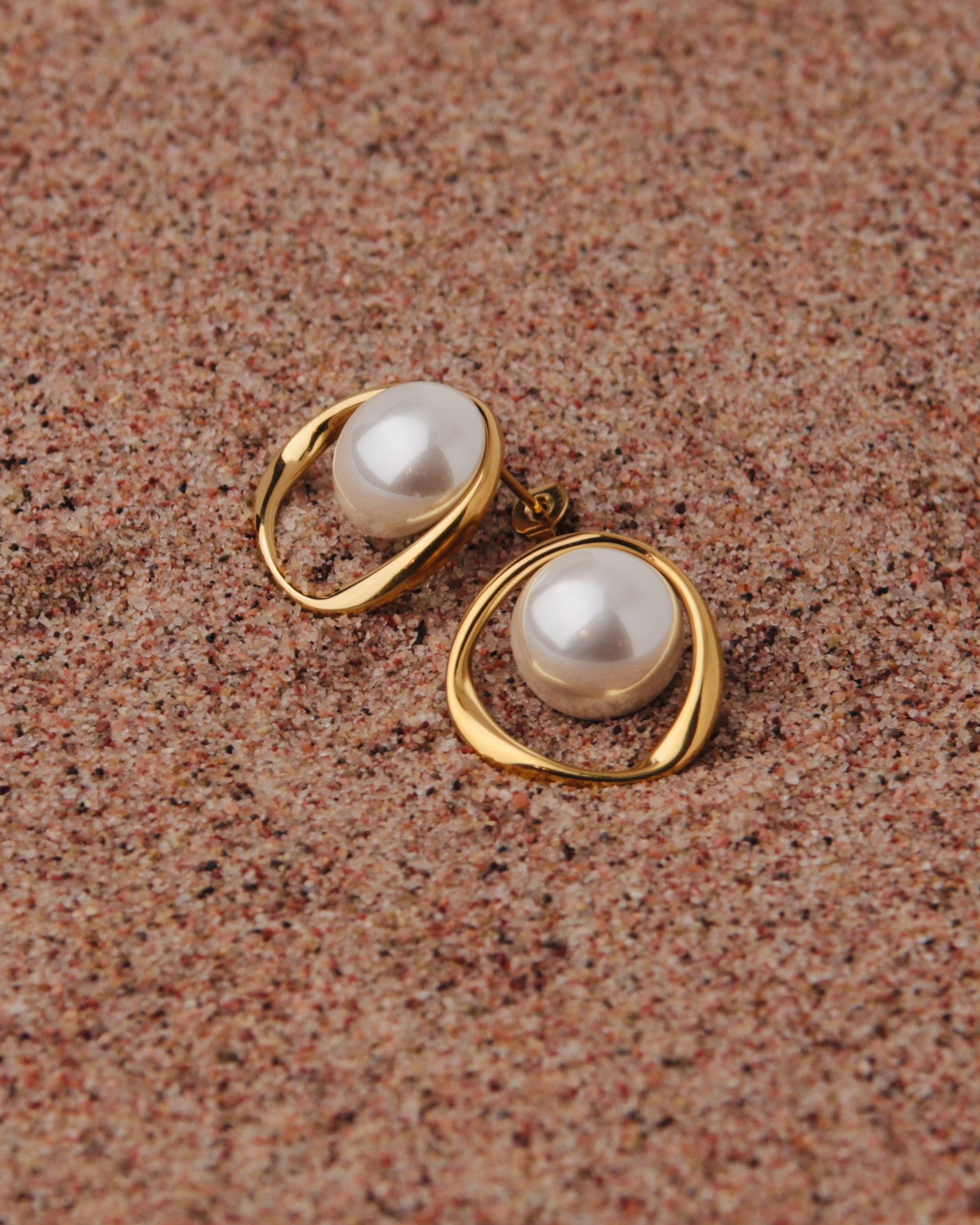 A pair of gold-toned statement earrings with large white freshwater pearls, displayed on a textured brown sand background.