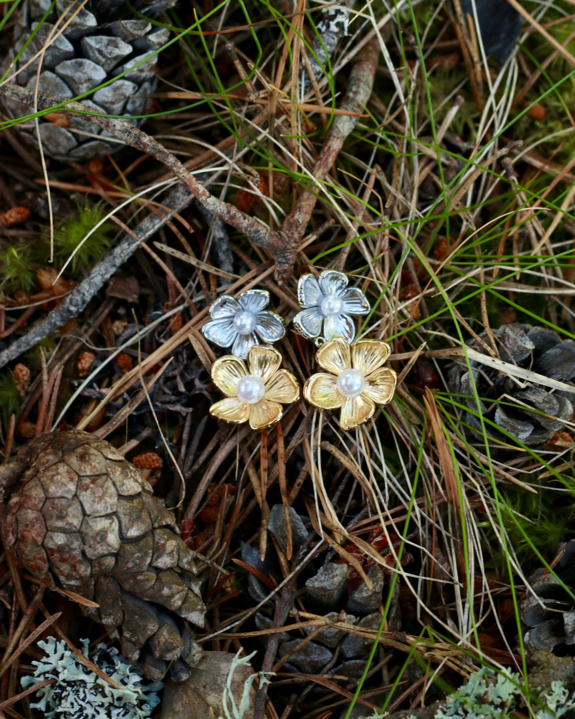 A pair of flower-shaped earrings placed on forest floor moss and pine needles. One earring is silver-toned with a pearl center, and the other is gold-toned with a pearl center.