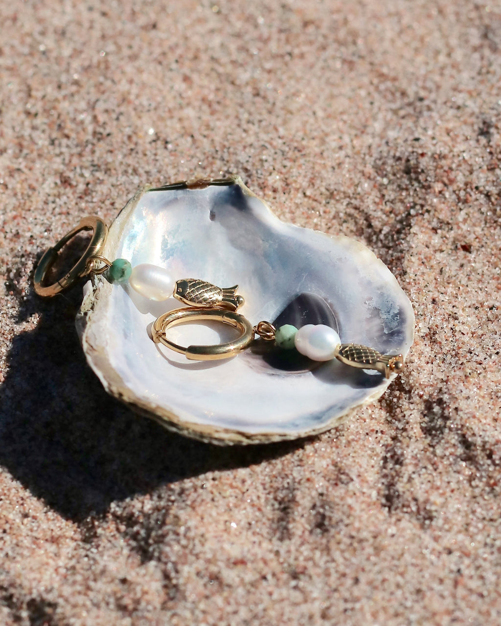 Gold hoop earrings with freshwater pearl and fish charms displayed in a seashell on a sandy beach by HNKO Finland