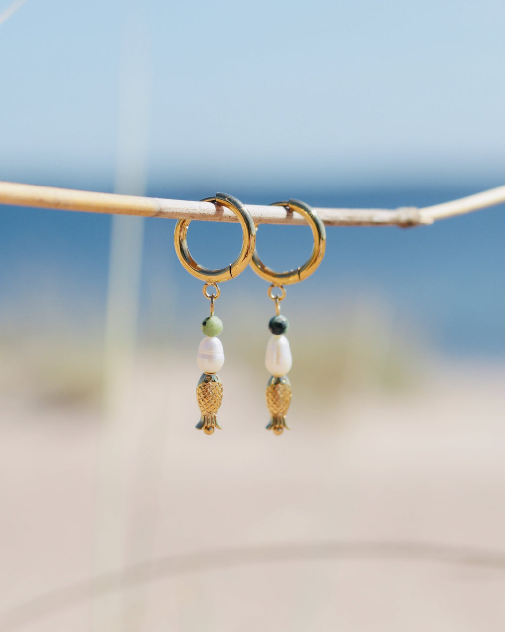 A pair of gold hoop earrings with dangling white beads, small green beads, and gold fish charms, hanging from a thin branch with a blurred beach background.