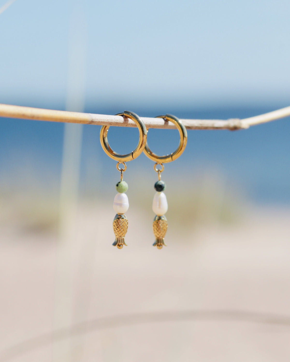 A pair of gold hoop earrings with dangling white beads, small green beads, and gold fish charms, hanging from a thin branch with a blurred beach background.