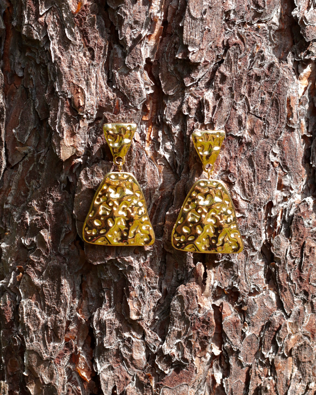 A pair of textured gold drop earrings with triangular puffy charms, displayed on a textured brown tree bark background.