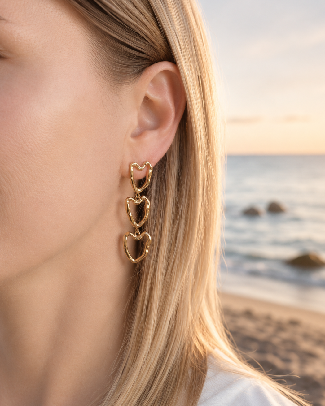 Gold heart-shaped earrings worn by a person with a beach background