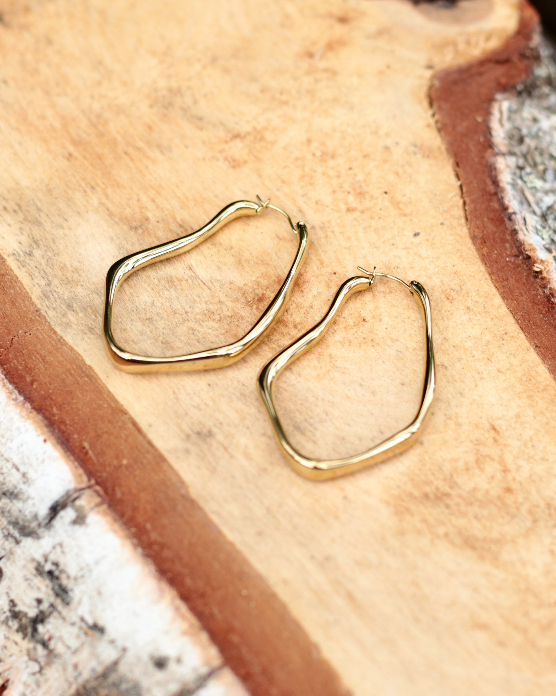 A pair of gold-toned abstract open hoop earrings resting on a textured, warm tan surface.