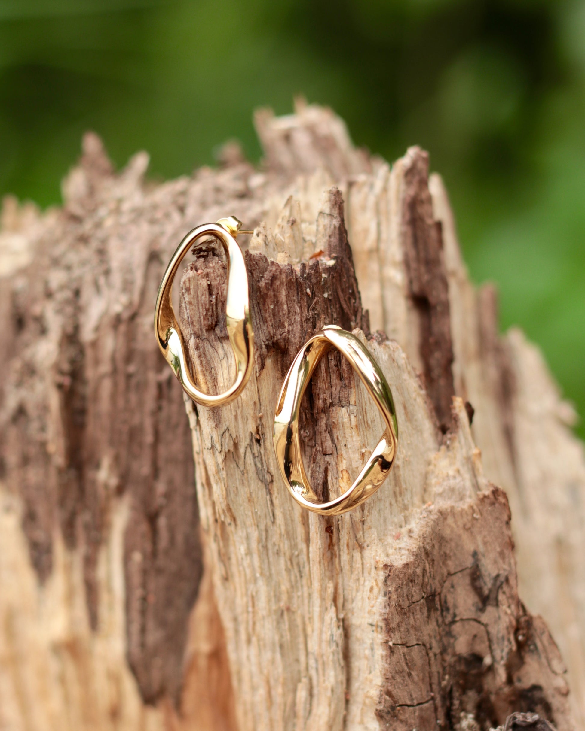 Two gold-toned abstract hoop earrings displayed on a textured piece of weathered wood with a blurred green outdoor background.