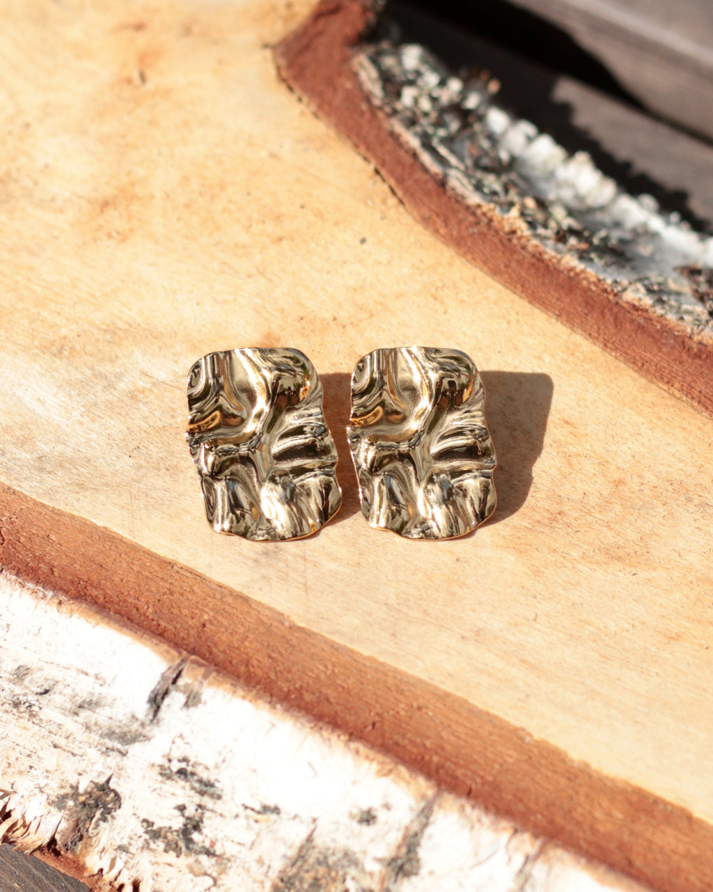 A pair of chunky, textured gold-toned stud earrings resting on a rustic wood slice background.