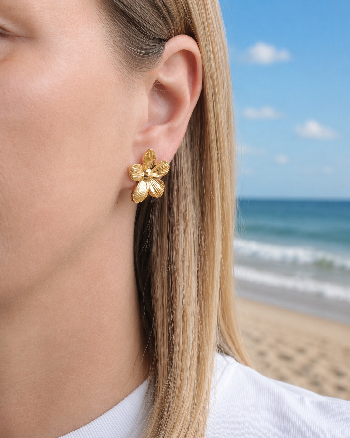 Gold flower-shaped earring worn by a person with a beach in the background