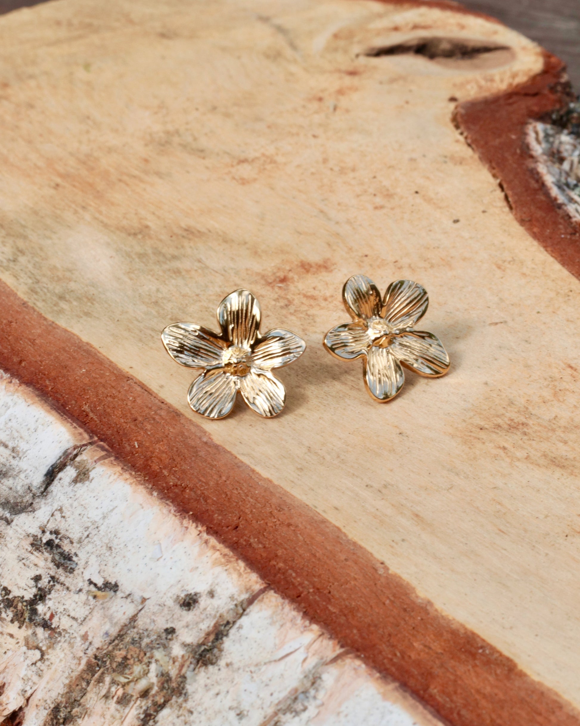 A pair of small gold flower-shaped stud earrings resting on a natural wood slice background.