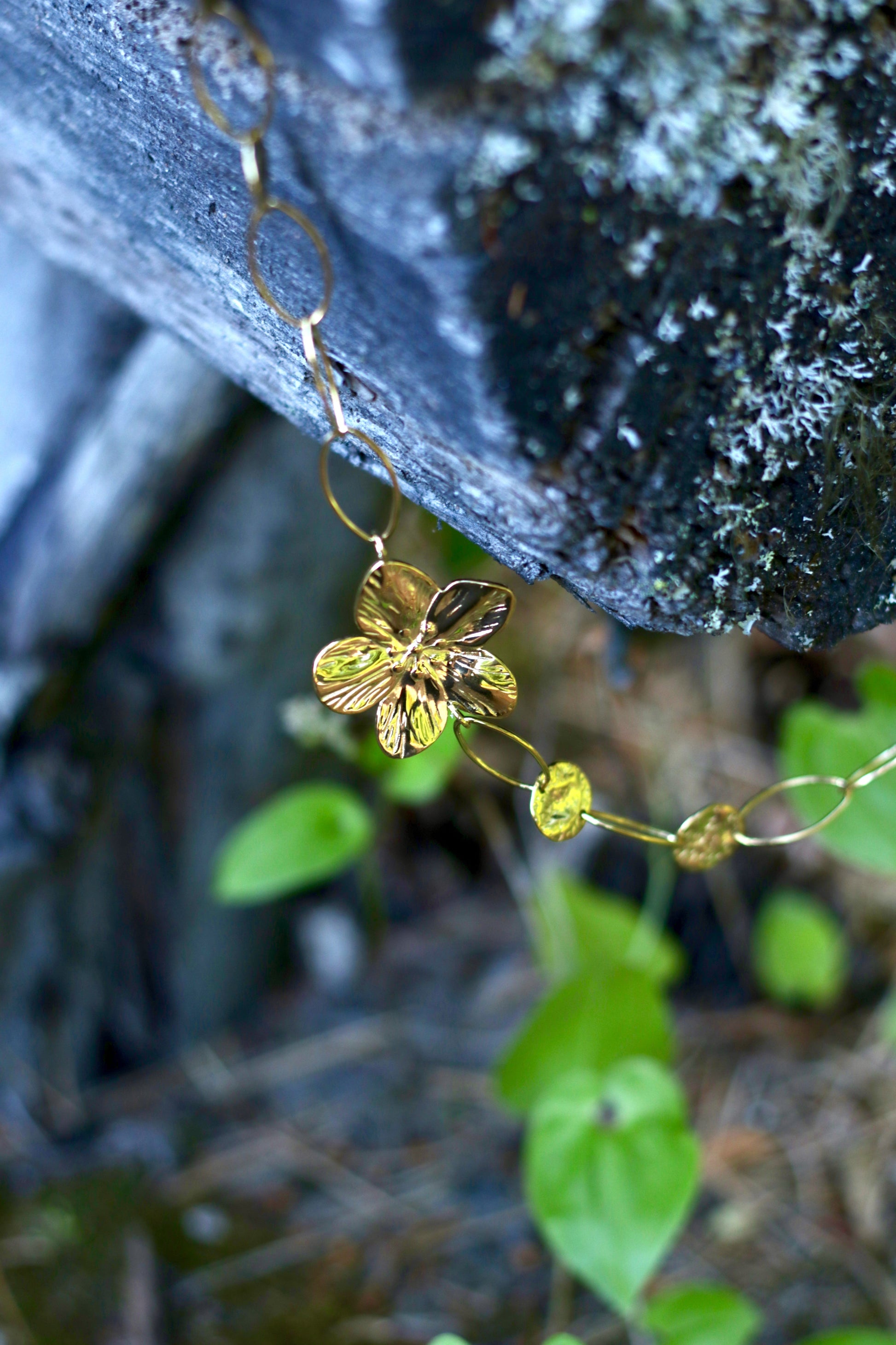 A close-up of a gold-toned flower-shaped chain necklace draped over a dark textured rock with green leaves blurred in the background.