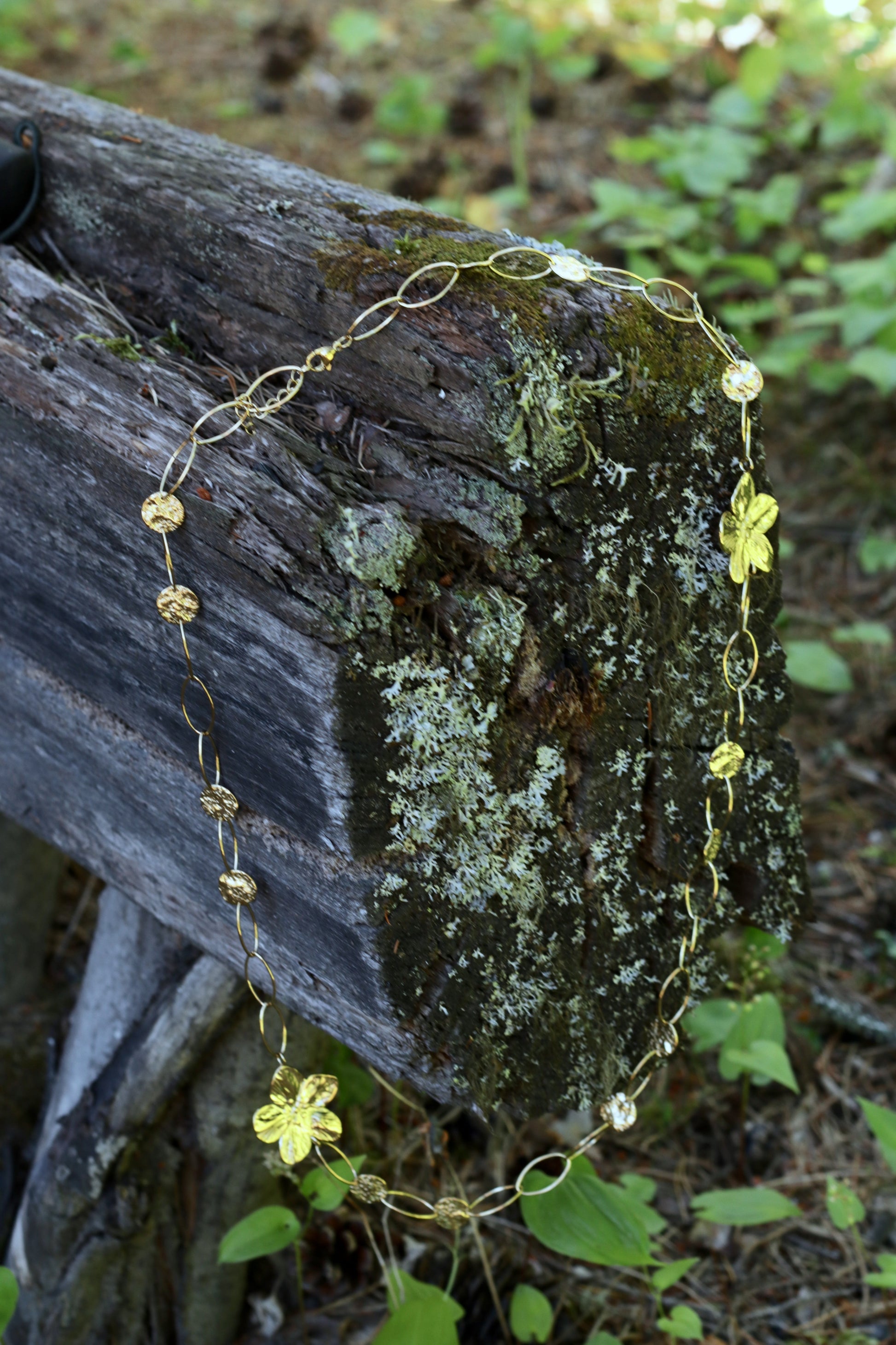 A delicate gold chain necklace with small circular medallions and flower charms draped over a mossy log outdoors, with green foliage blurred in the background.
