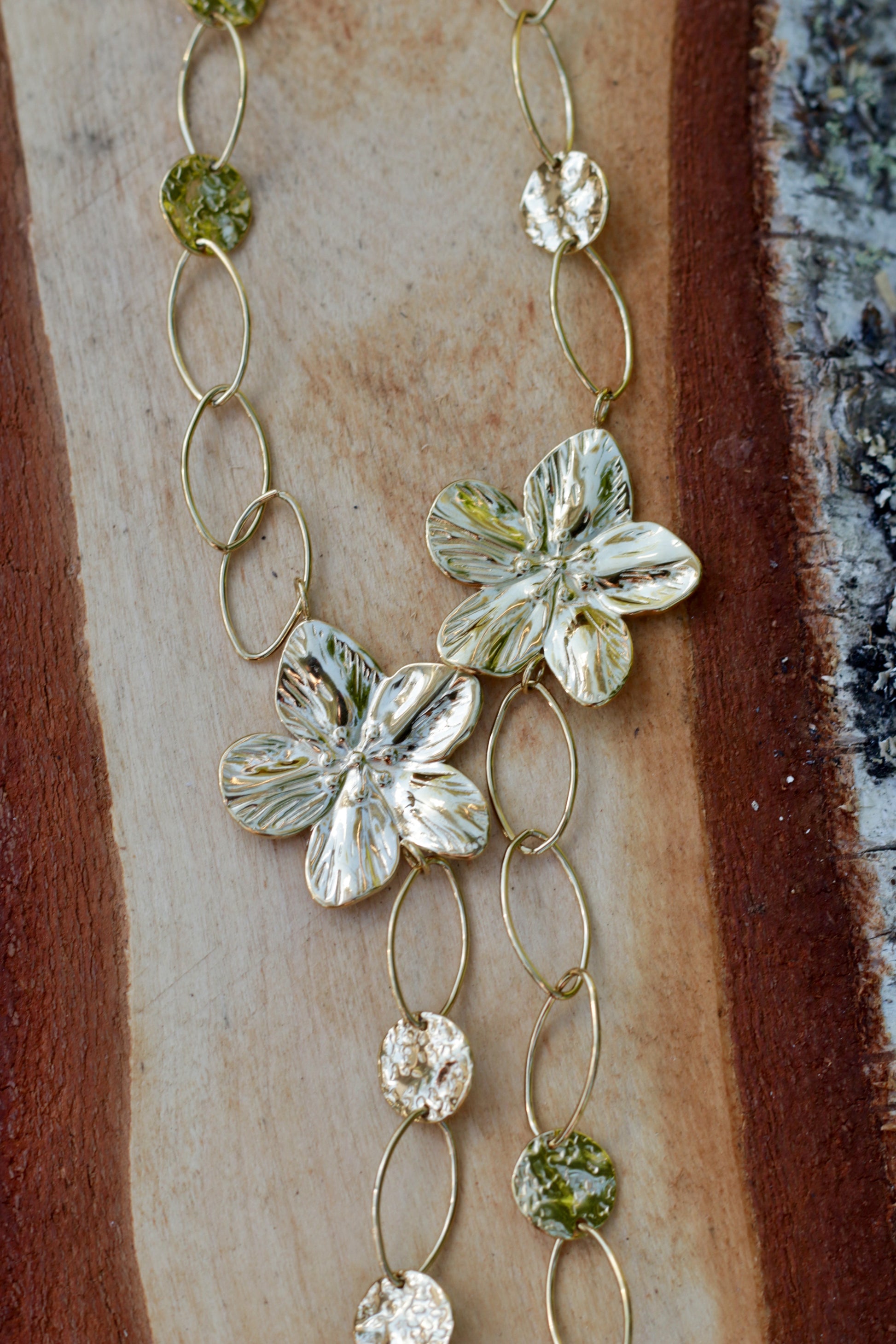 A close-up of a gold-toned necklace featuring textured silver flower charms and faceted green and clear beads, laid on a wood slice background.