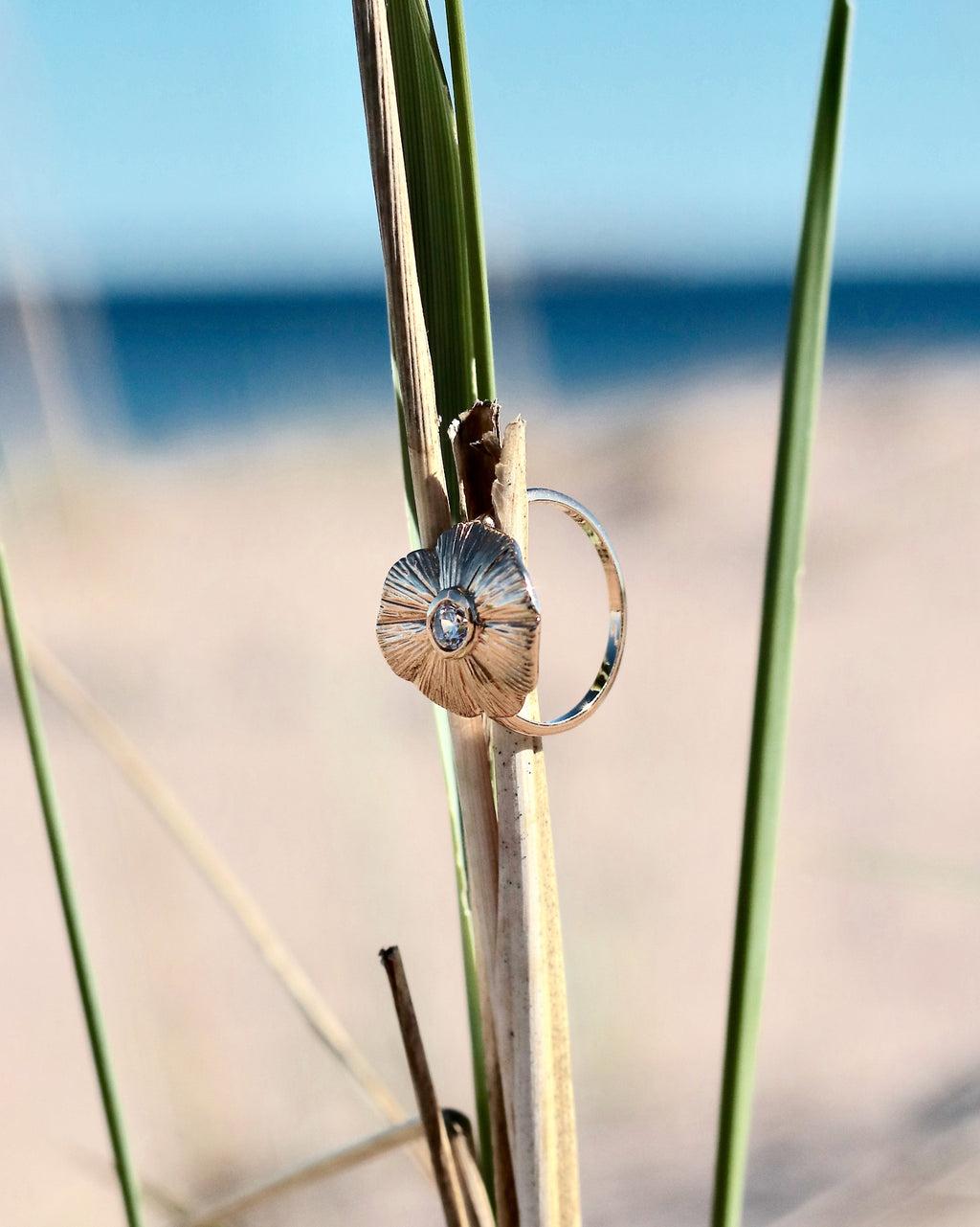 18K gold-plated floral ring with sculpted petals and a luminous center stone displayed on coastal vegetation in Hanko, Finland