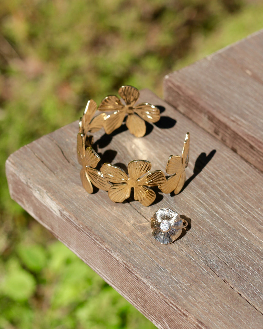 A gold-tone flower-shaped cuff bracelet and a matching flower stud earring displayed on a weathered wooden board with a blurred green outdoor background.