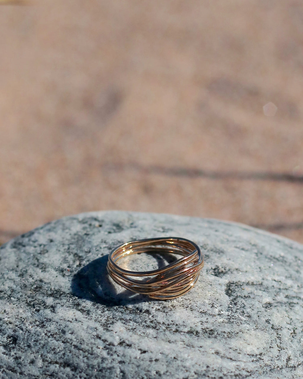 18K gold-plated interwoven band ring with flowing sculptural lines displayed on coastal stones at the beach in Hanko, Finland