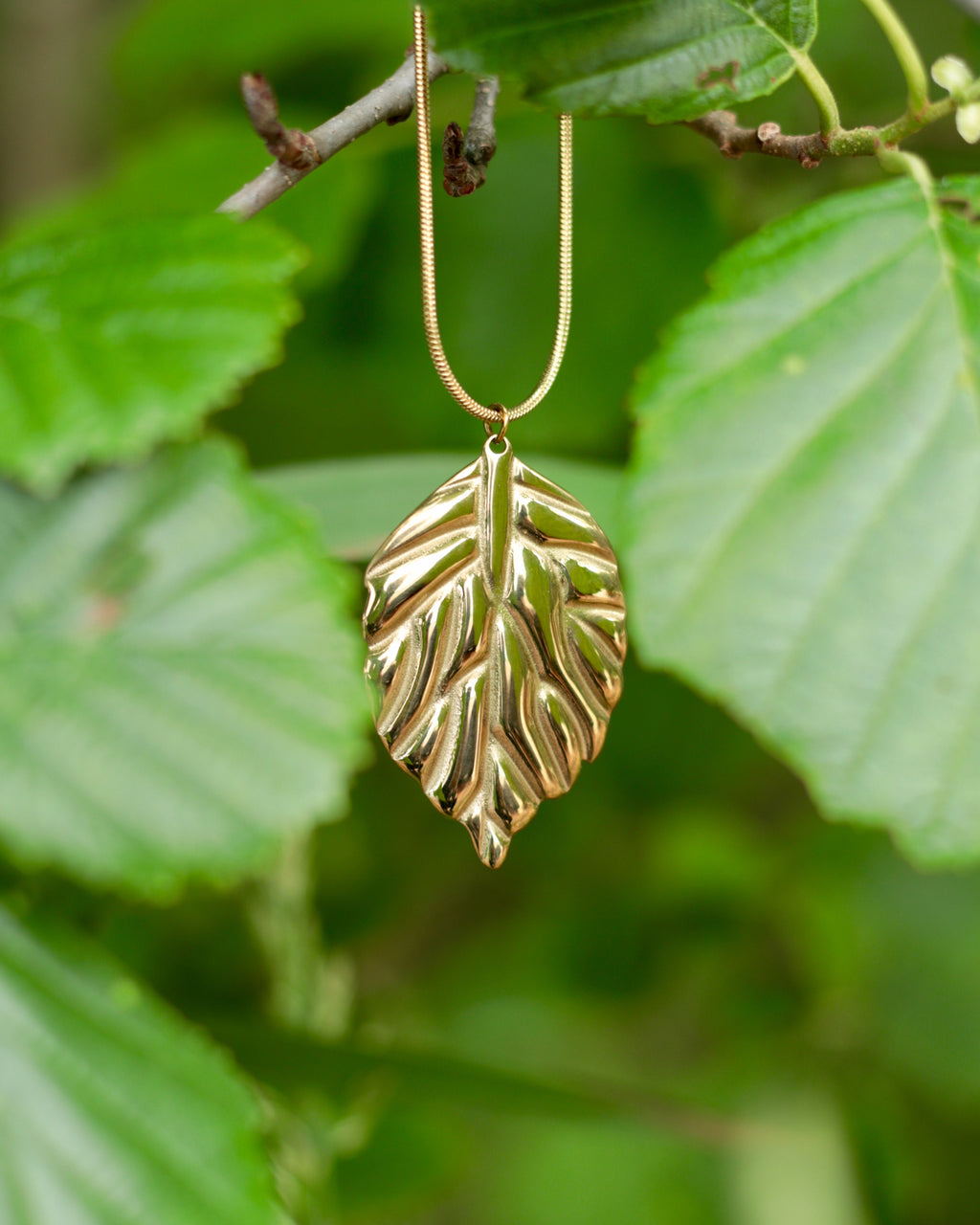 A gold leaf pendant necklace hanging from a thin gold chain, set against a soft-focus background of green leaves.