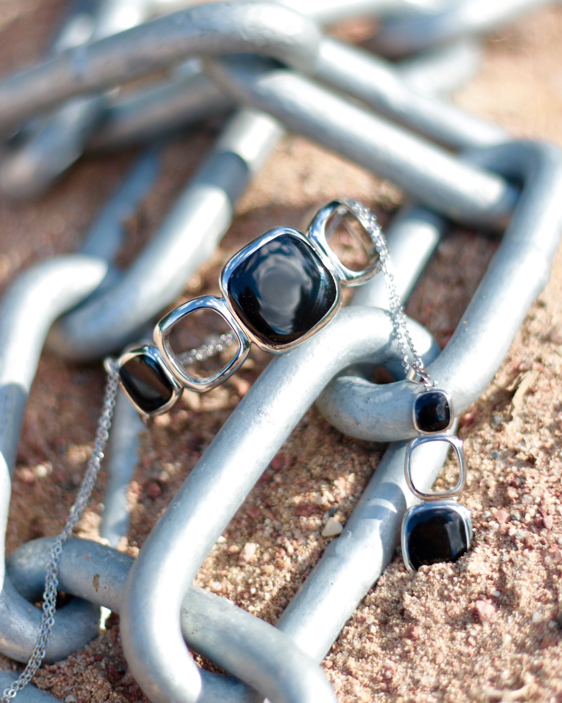 A silver-toned jewelry set featuring a chunky cuff bracelet and a matching drop earring, both adorned with black cabochon stones, displayed on a rustic chain over sandy ground.