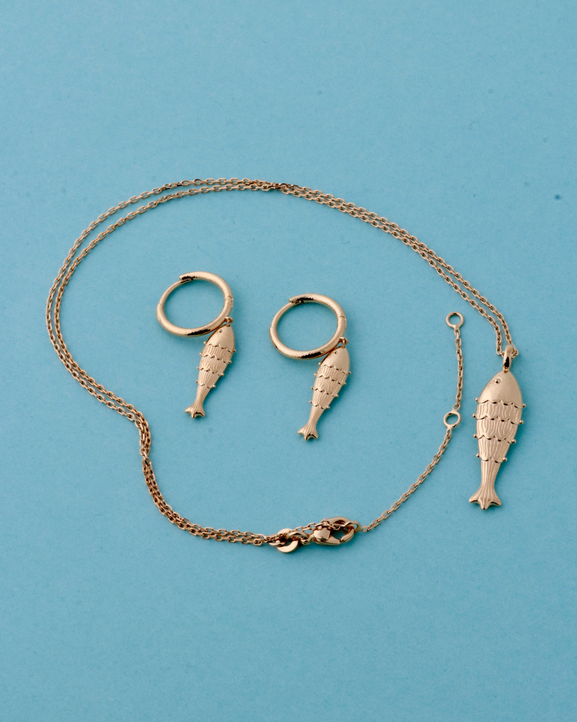 A gold jewelry set featuring a fish-shaped pendant necklace, matching fish-shaped hoop earrings, and a lobster clasp, displayed on a solid light blue background.