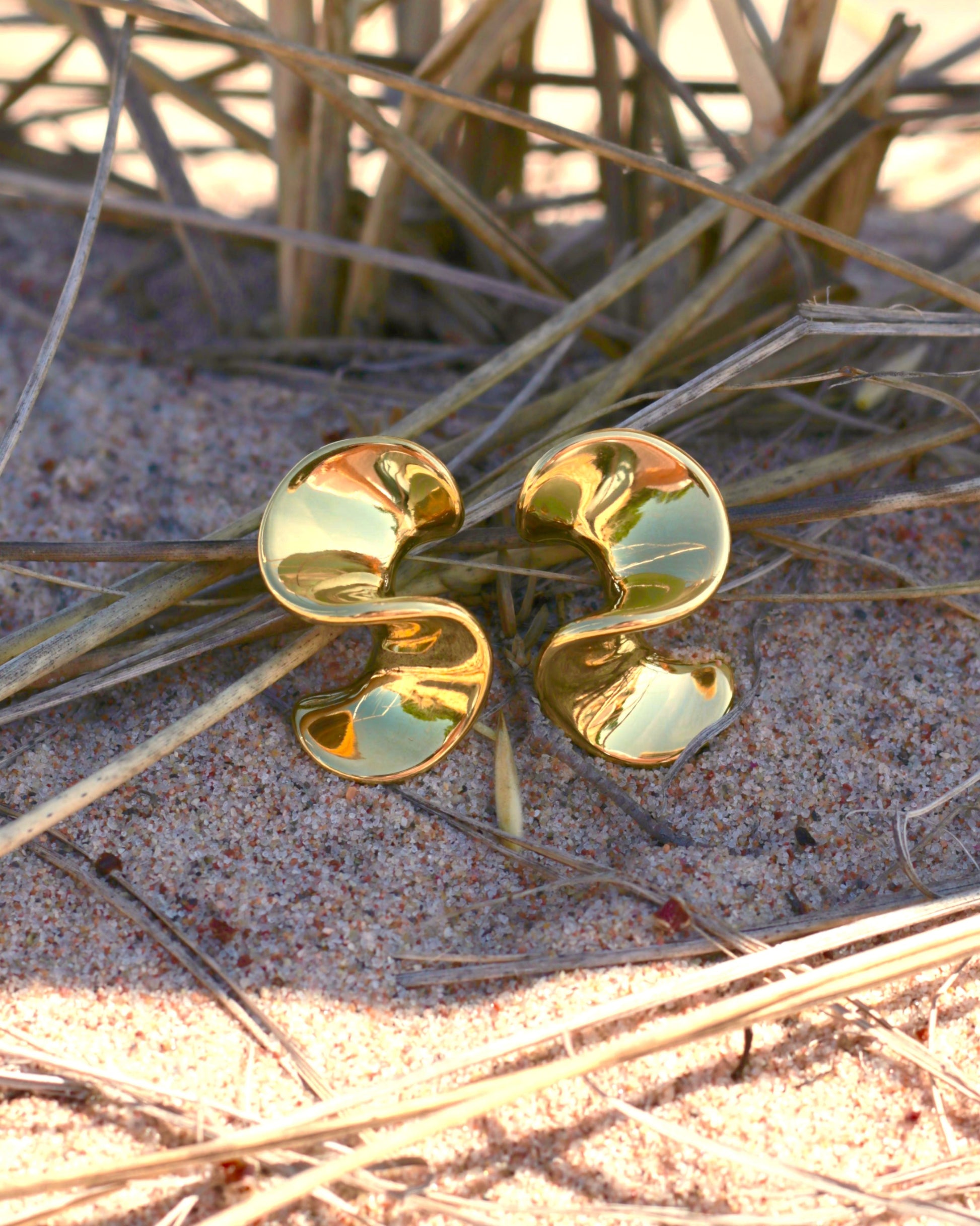 A pair of gold-toned, abstract spiral-shaped earrings resting on sand amidst dry grass.