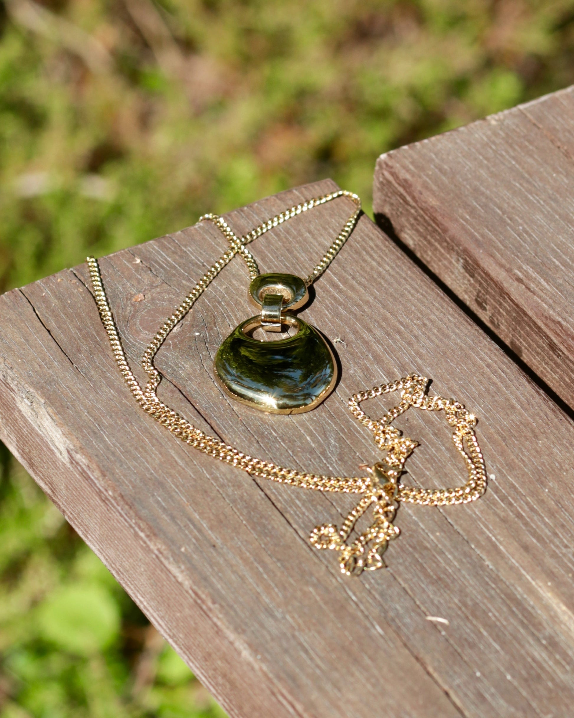 A gold chain necklace with a large, irregularly shaped dark green polished stone pendant, resting on weathered wooden planks with a blurred green outdoor background.