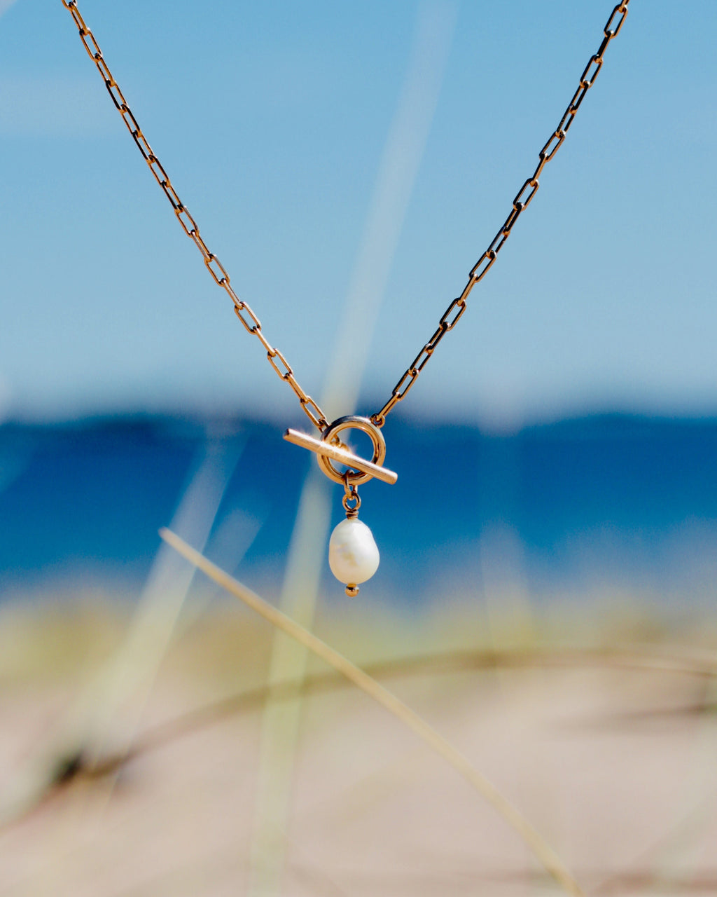 A delicate gold chain necklace with a toggle clasp and a single white pearl pendant, photographed outdoors against a blurred beach background with blue sky and sand.